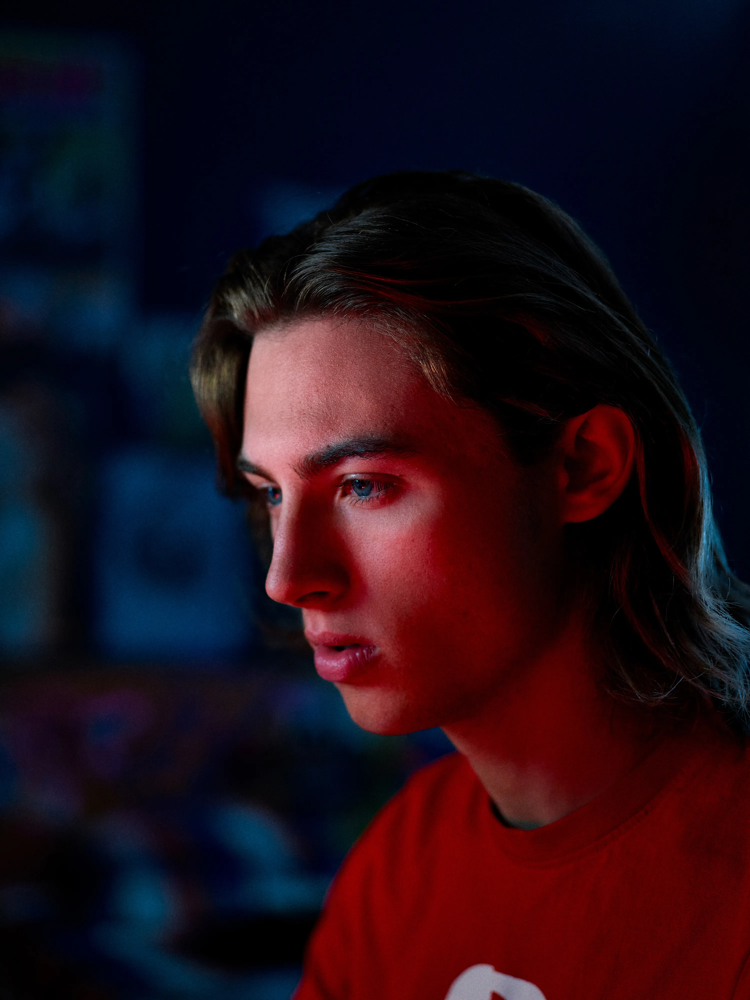Young man with long hair wearing a red shirt, illuminated by red and blue lighting, staring thoughtfully to the left against a dark, blurred background.