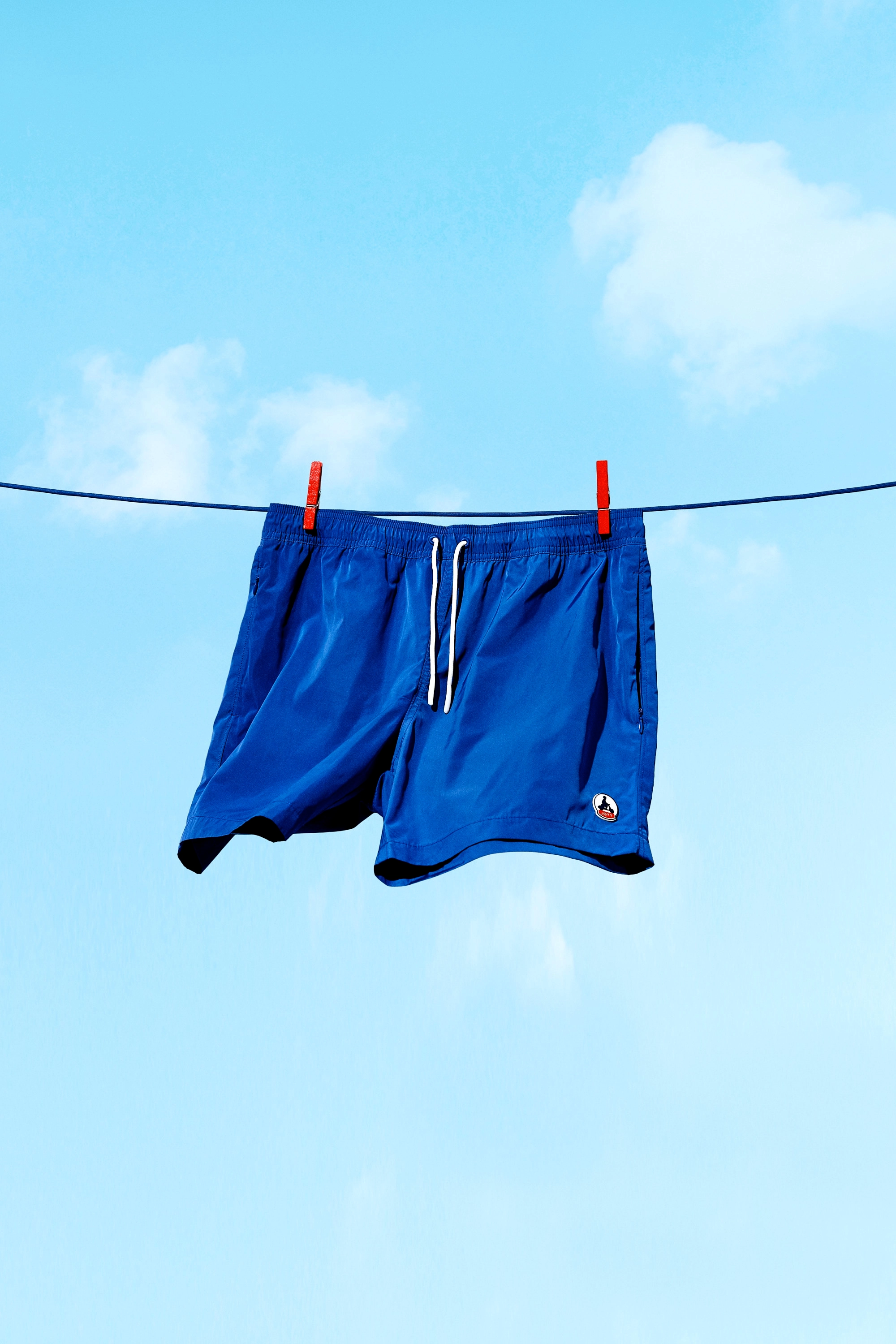 Blue swim shorts hanging on a clothesline with red clothespins against a blue sky with scattered clouds.