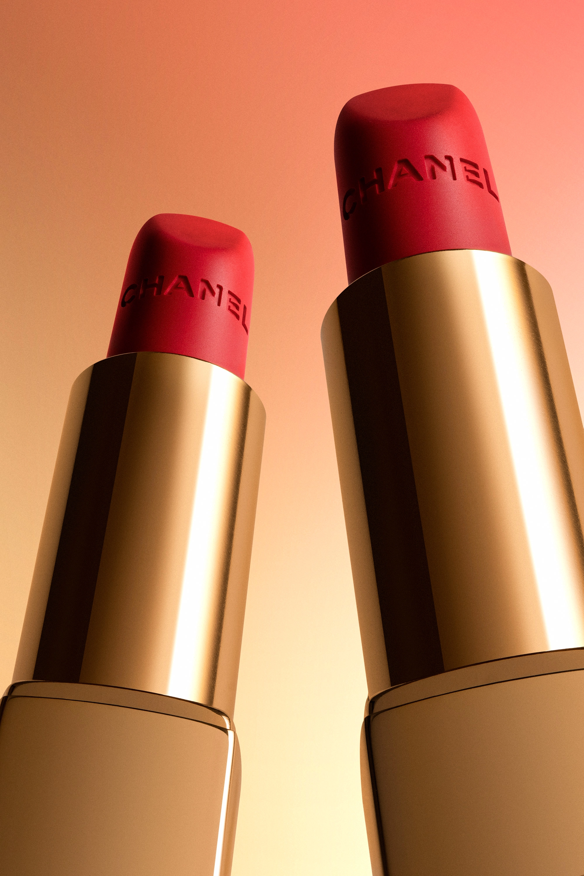 Close-up of two gold Chanel lipsticks with red tips embossed with 'CHANEL' lettering, against a gradient peach background.