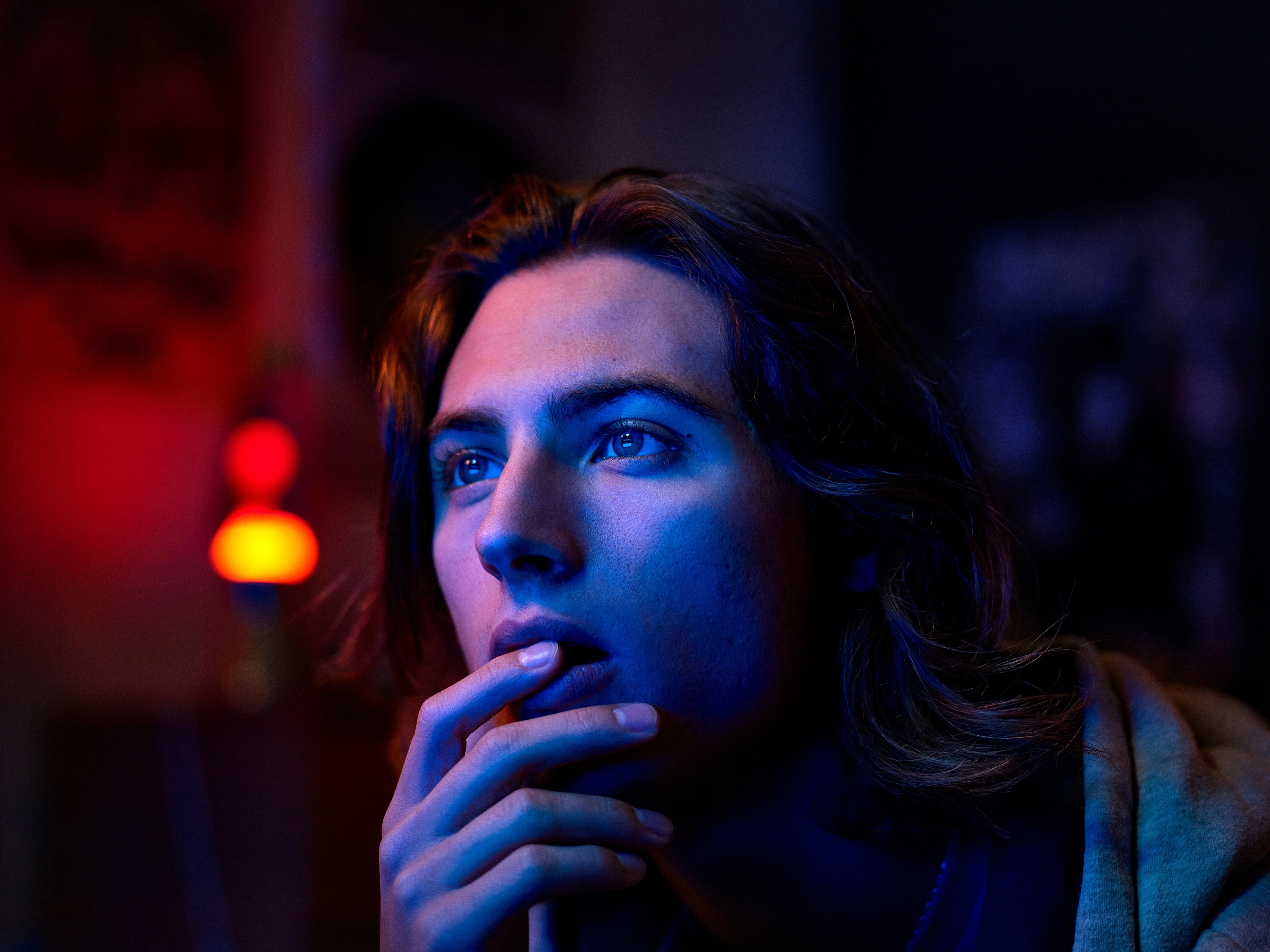 Close-up of a young person with long hair illuminated by red and blue lighting, looking thoughtfully to the side.