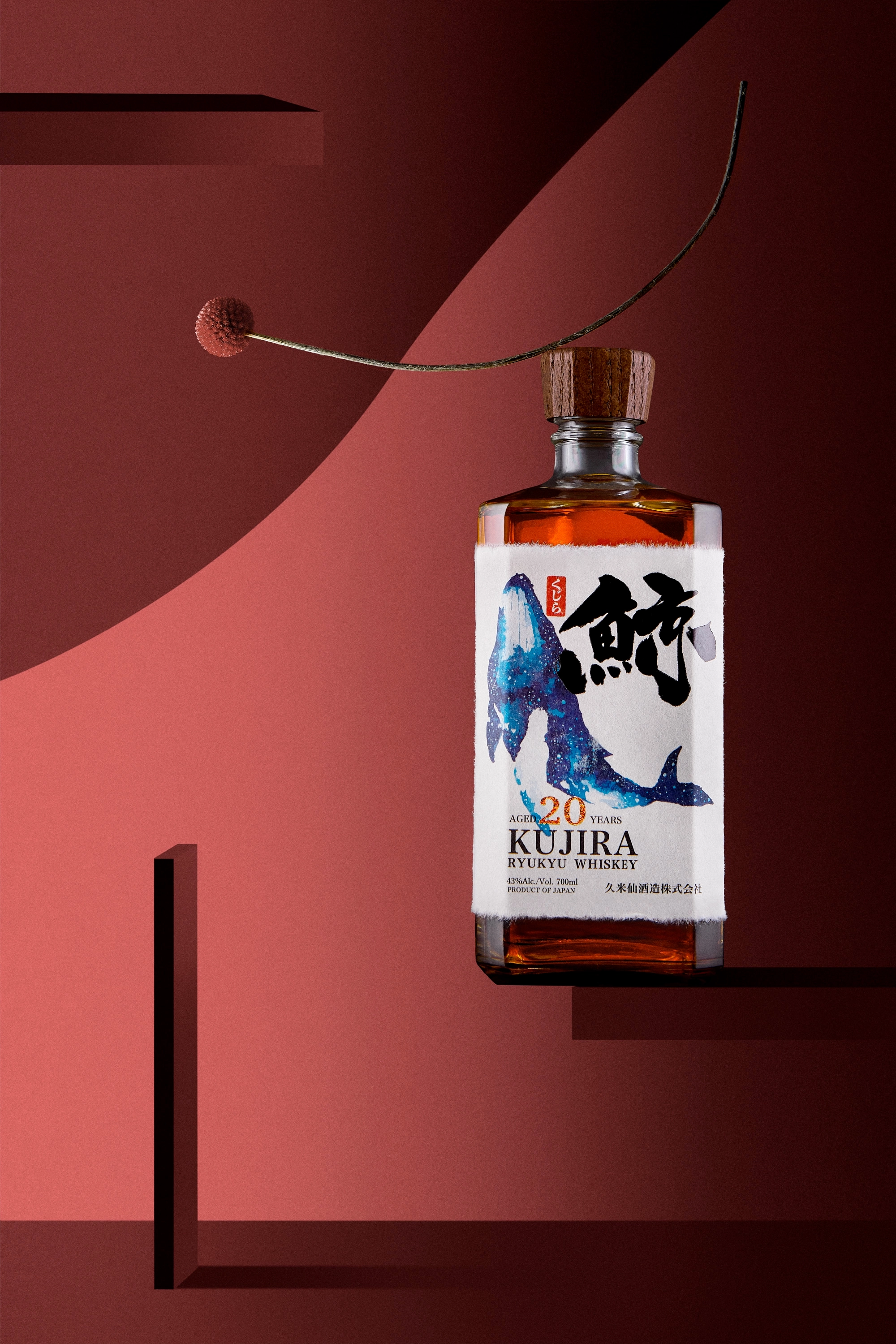 Bottle of Kujira 20-year aged Ryukyu whiskey with a wooden cap and a label featuring a whale illustration, set against a red geometric background with a long curved twig and round seed.