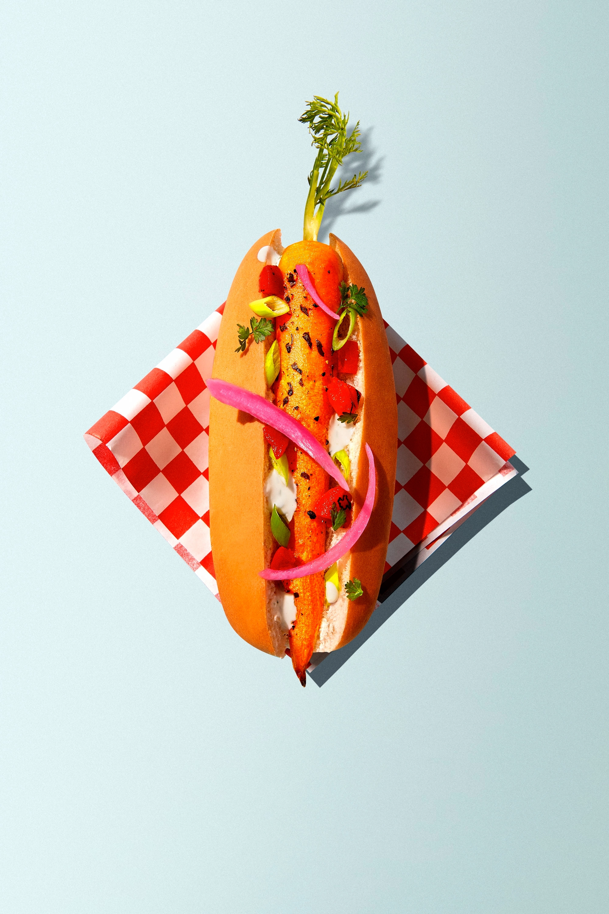 Carrot hot dog with grilled carrot, pickled onions, herbs, and sauce in a bun on a red and white checkered napkin.