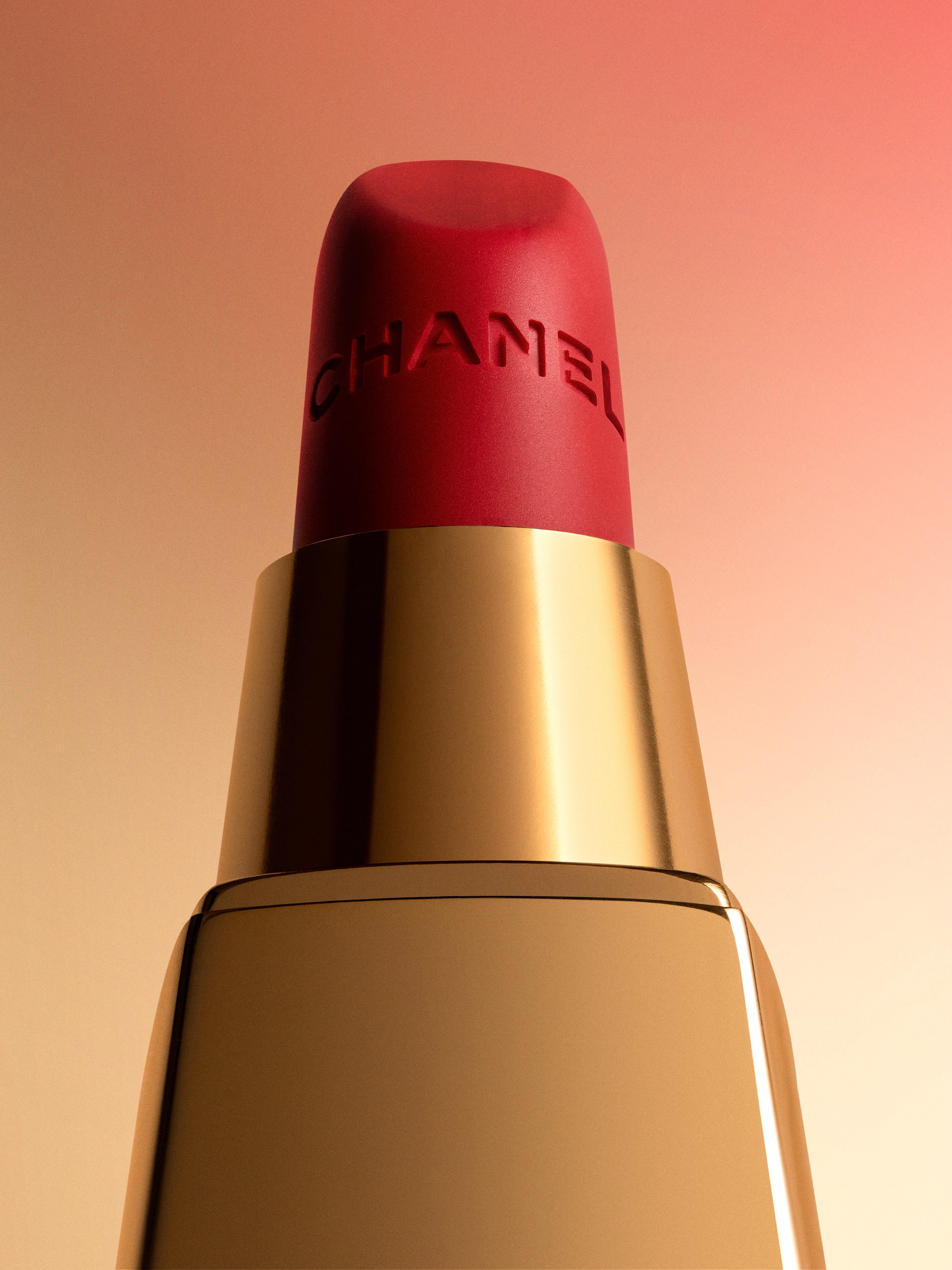 Close-up of red Chanel lipstick with gold casing on a pink and beige gradient background.