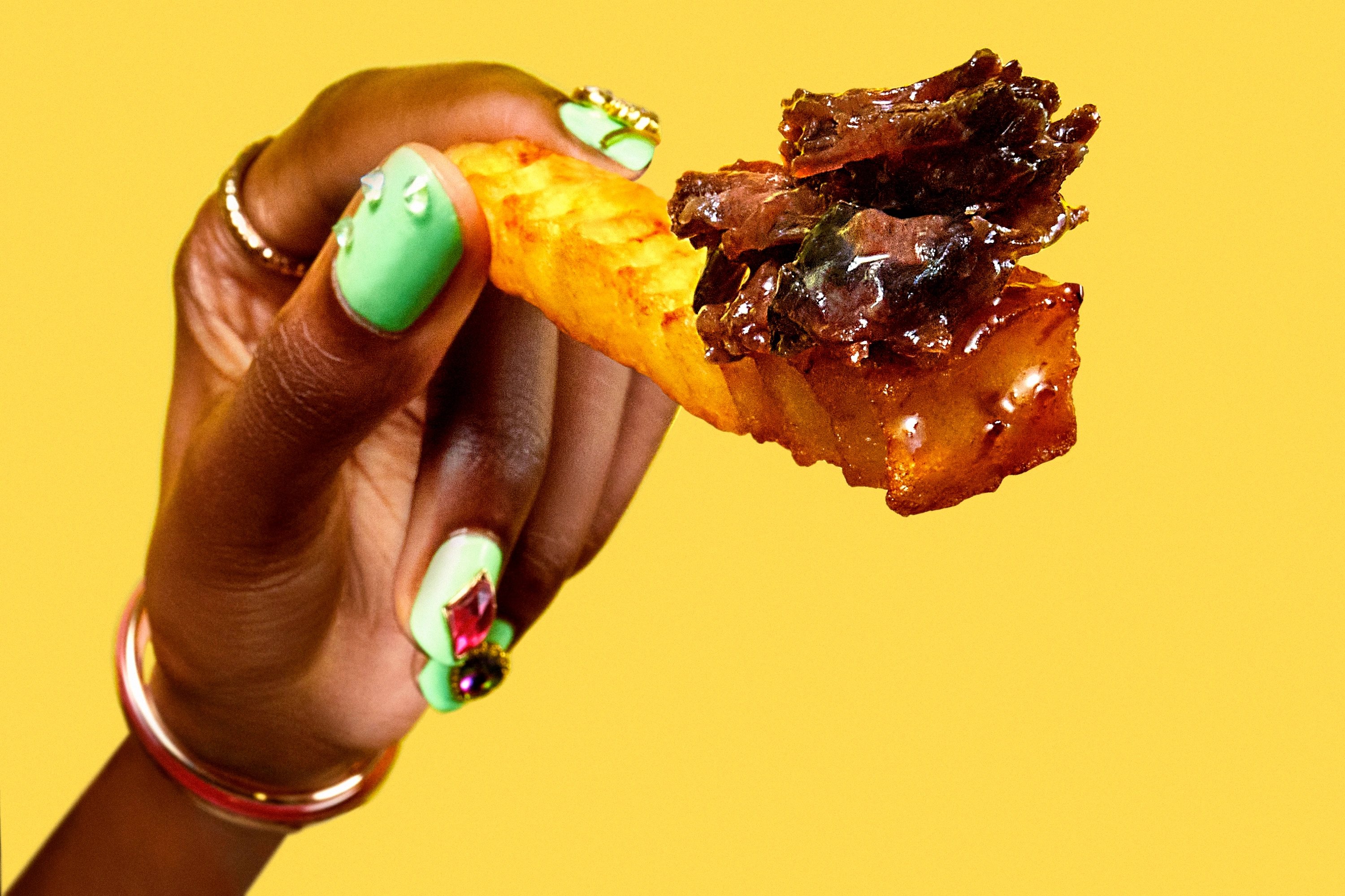 Hand with mint green nails holding a crinkle-cut fry topped with glazed beef pieces against a yellow background.