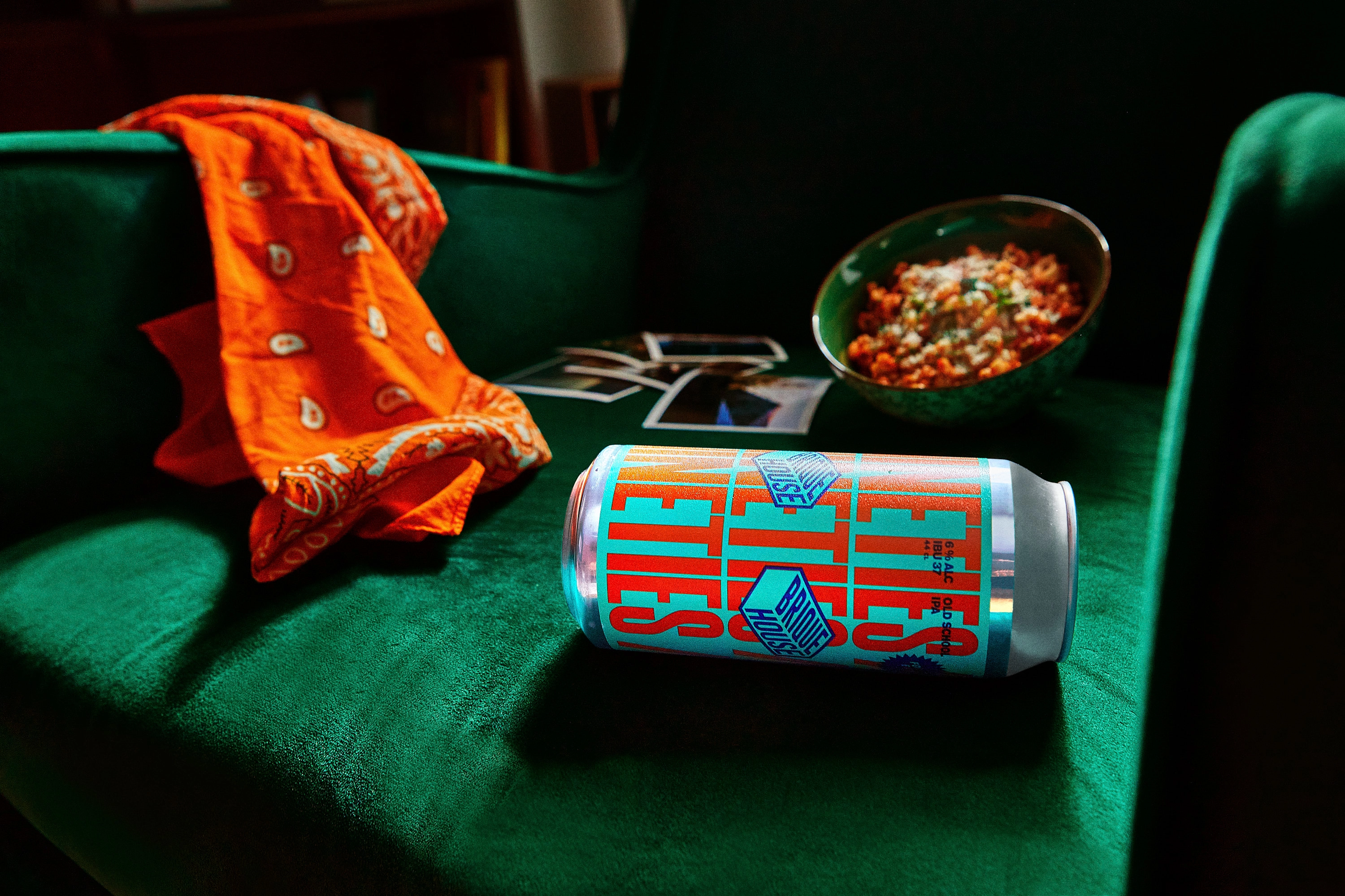 Green velvet sofa with an orange bandana draped over the armrest, a can of beer labeled 'Old School IPA' lying on its side, and a bowl of pasta with sauce in the background.