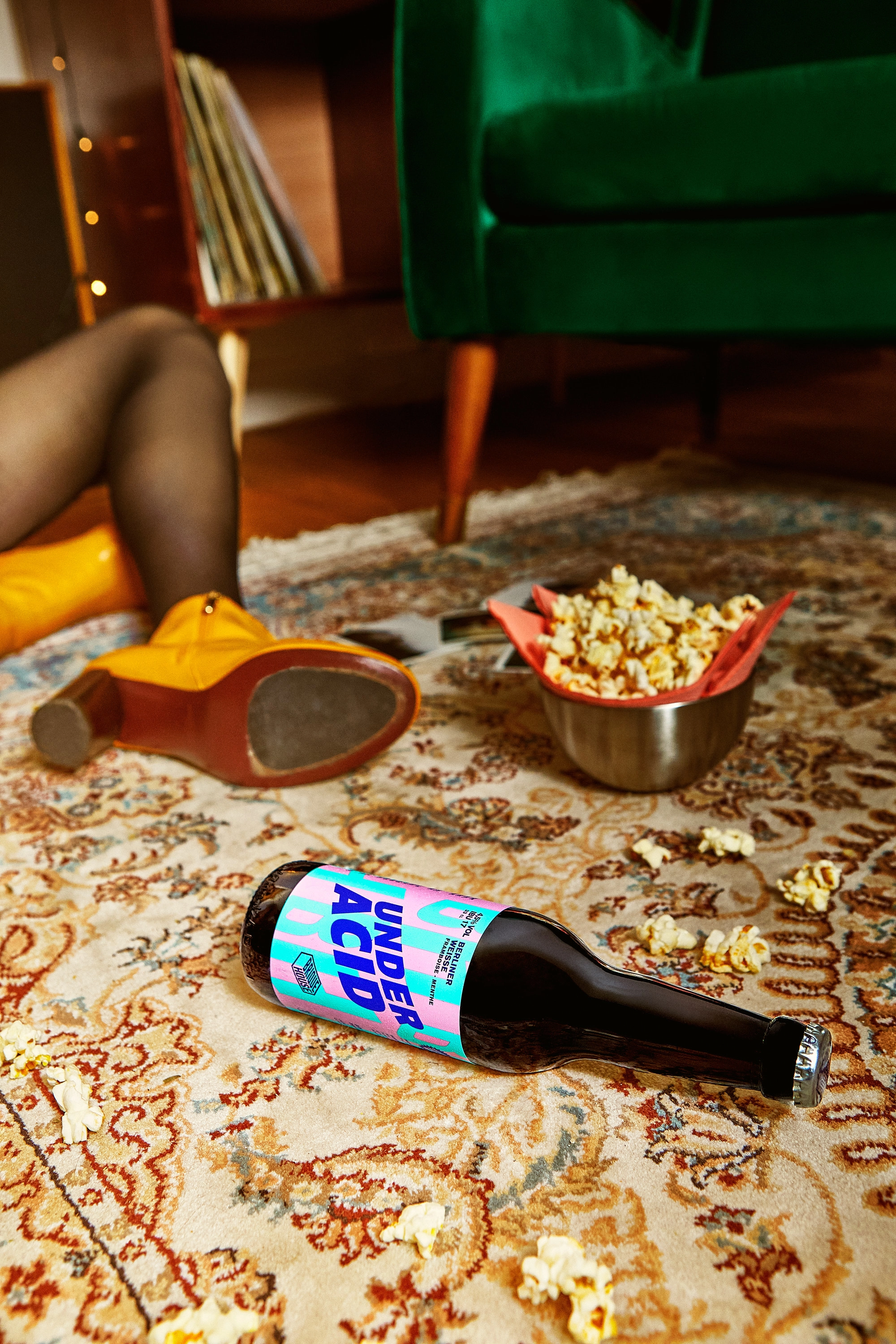 Bottle labeled 'Under Acid' lying on a patterned rug with popcorn scattered nearby and person wearing yellow boots sitting in the background.