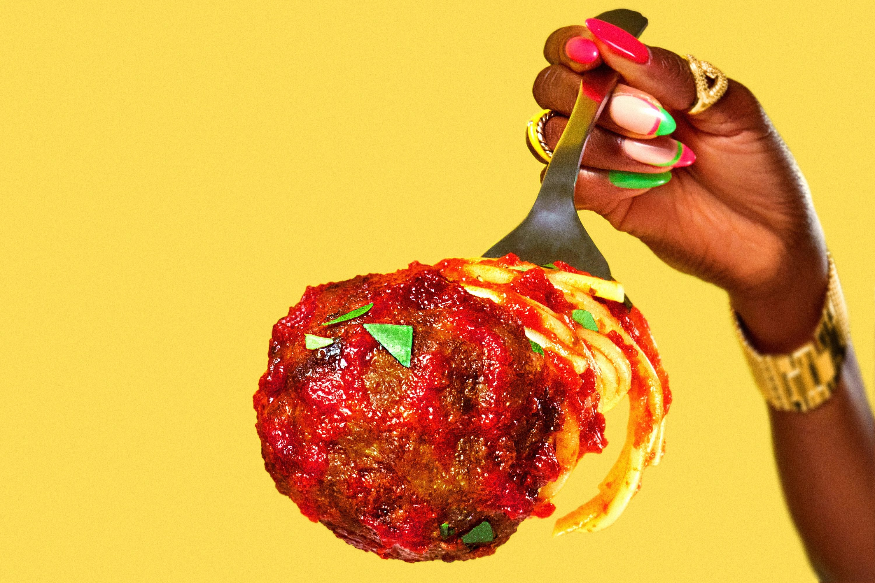 Hand with colorful nails holding a fork with spaghetti and a large meatball covered in red sauce and herbs against a yellow background.
