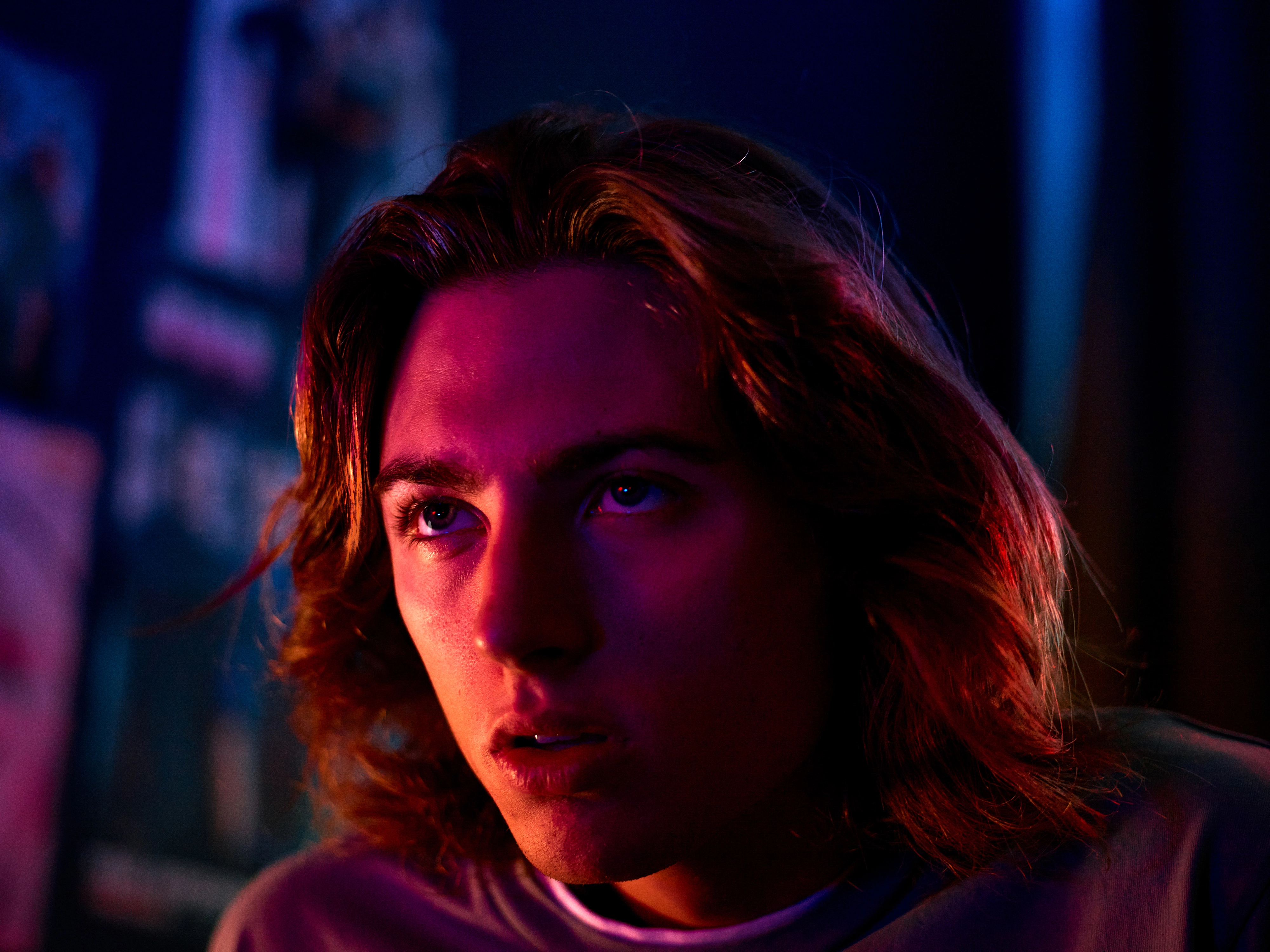 Young man with long hair illuminated by red and purple lighting looking upward thoughtfully in a dark environment.