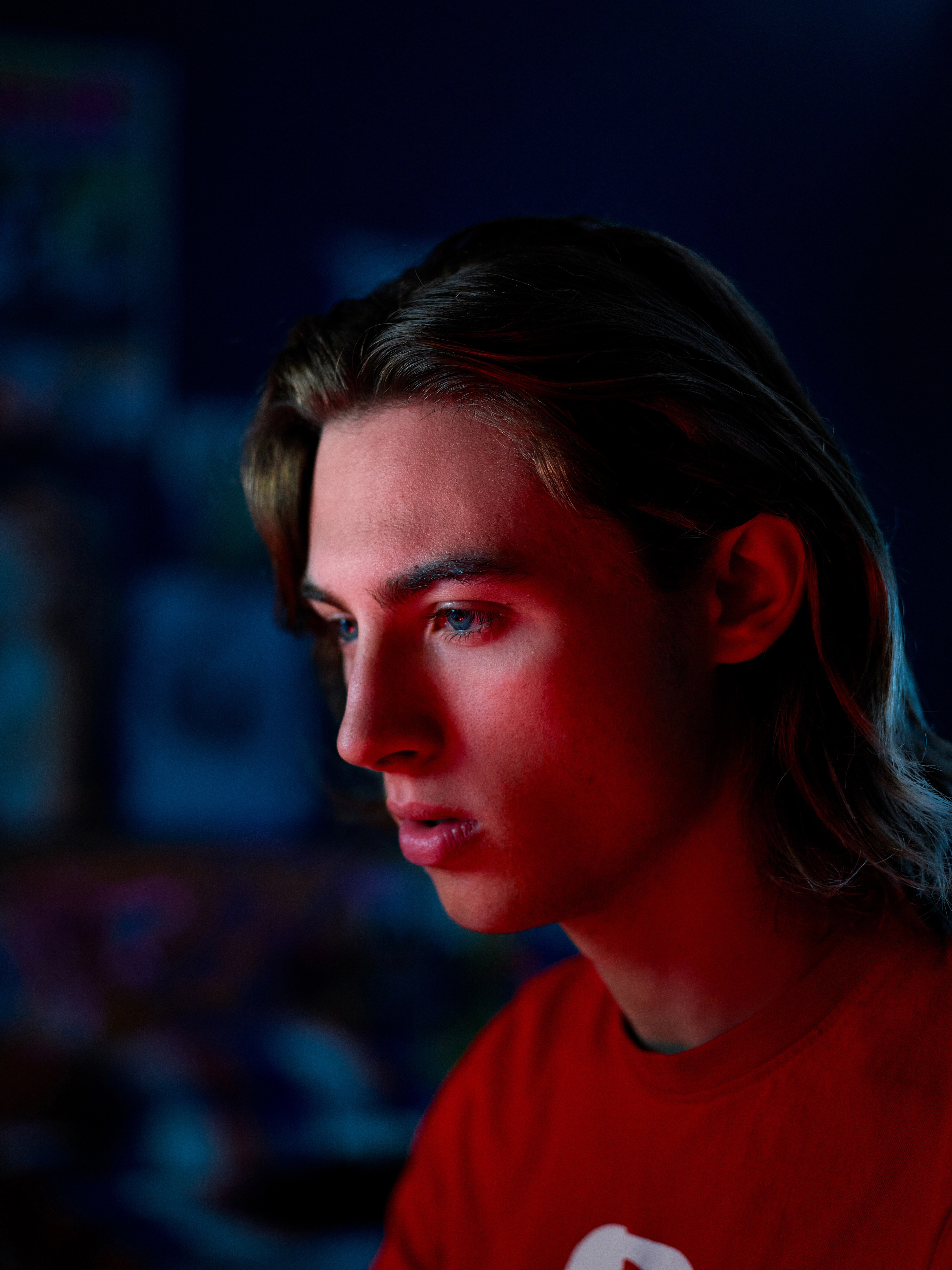 Young man with long hair and blue eyes lit by red and blue lighting, looking thoughtfully to the side.