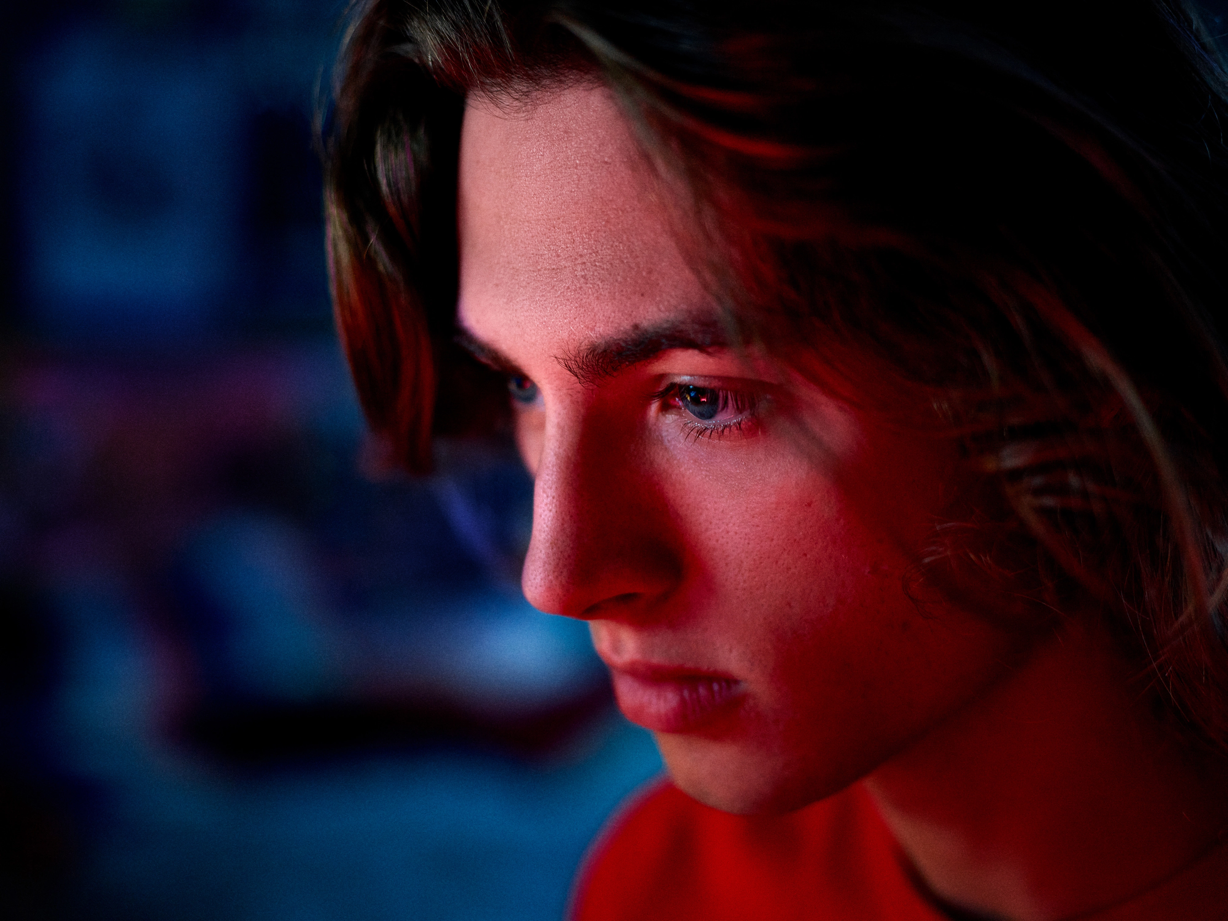 Close-up of a young person’s face illuminated by red and blue light, looking thoughtfully to the side.