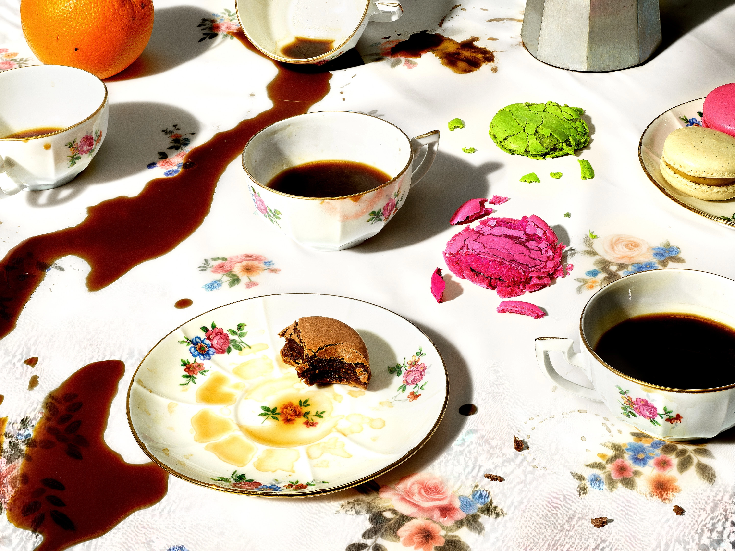 Spilled coffee on a floral tablecloth with vintage teacups and broken colorful macarons scattered around.