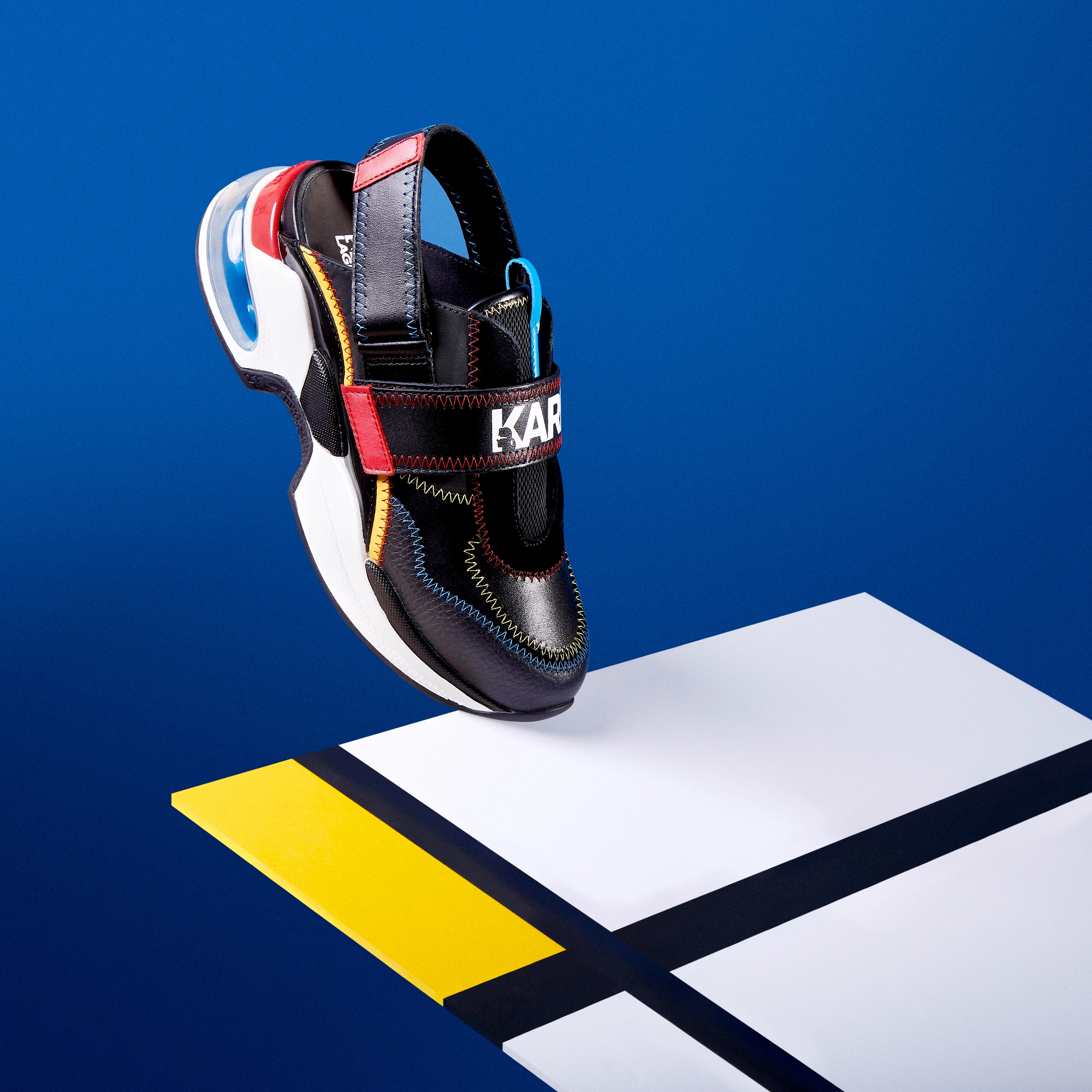 Black sandal with colorful stitching, red strap, and white thick sole floating above a white, yellow, and black geometric surface on a blue background.