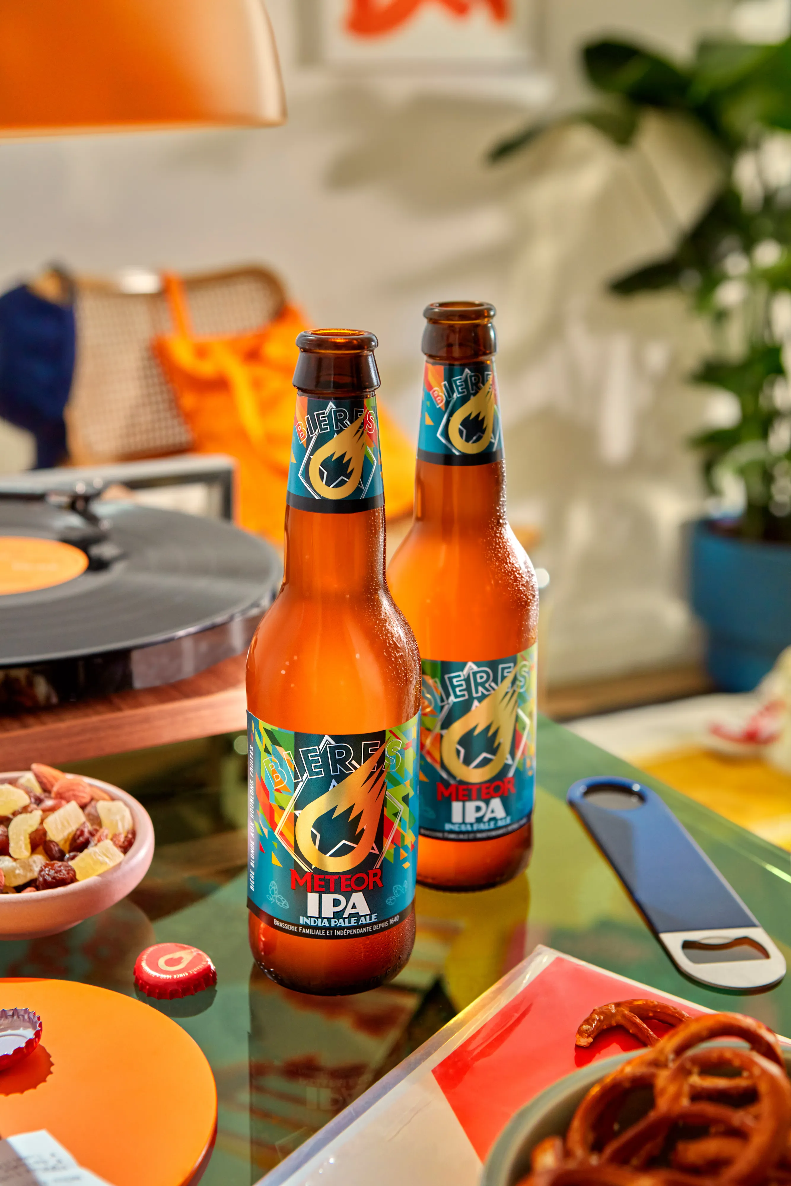 Two bottles of Meteor IPA beer on a glass table surrounded by snacks, a bottle opener, and a record player.