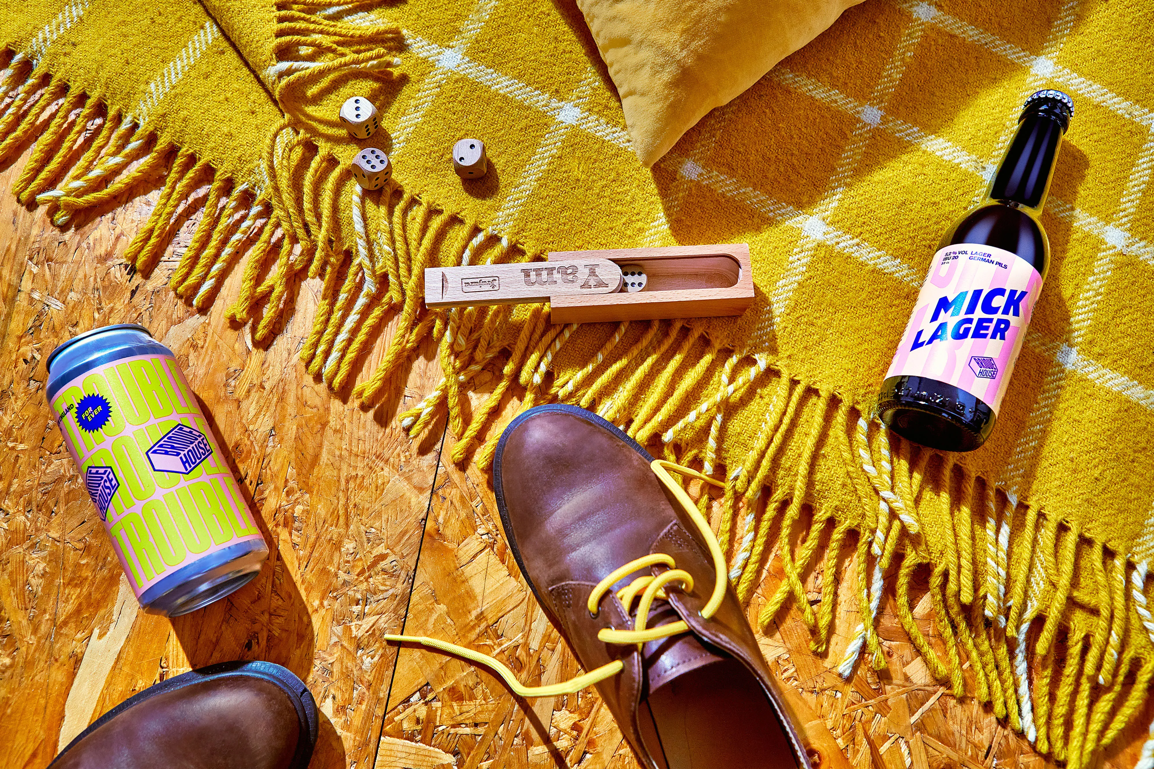 Brown leather shoe with yellow laces on wooden floor next to a yellow fringed blanket, dice in a wooden box, a can labeled Double Trouble, and a bottle labeled Mick Lager.