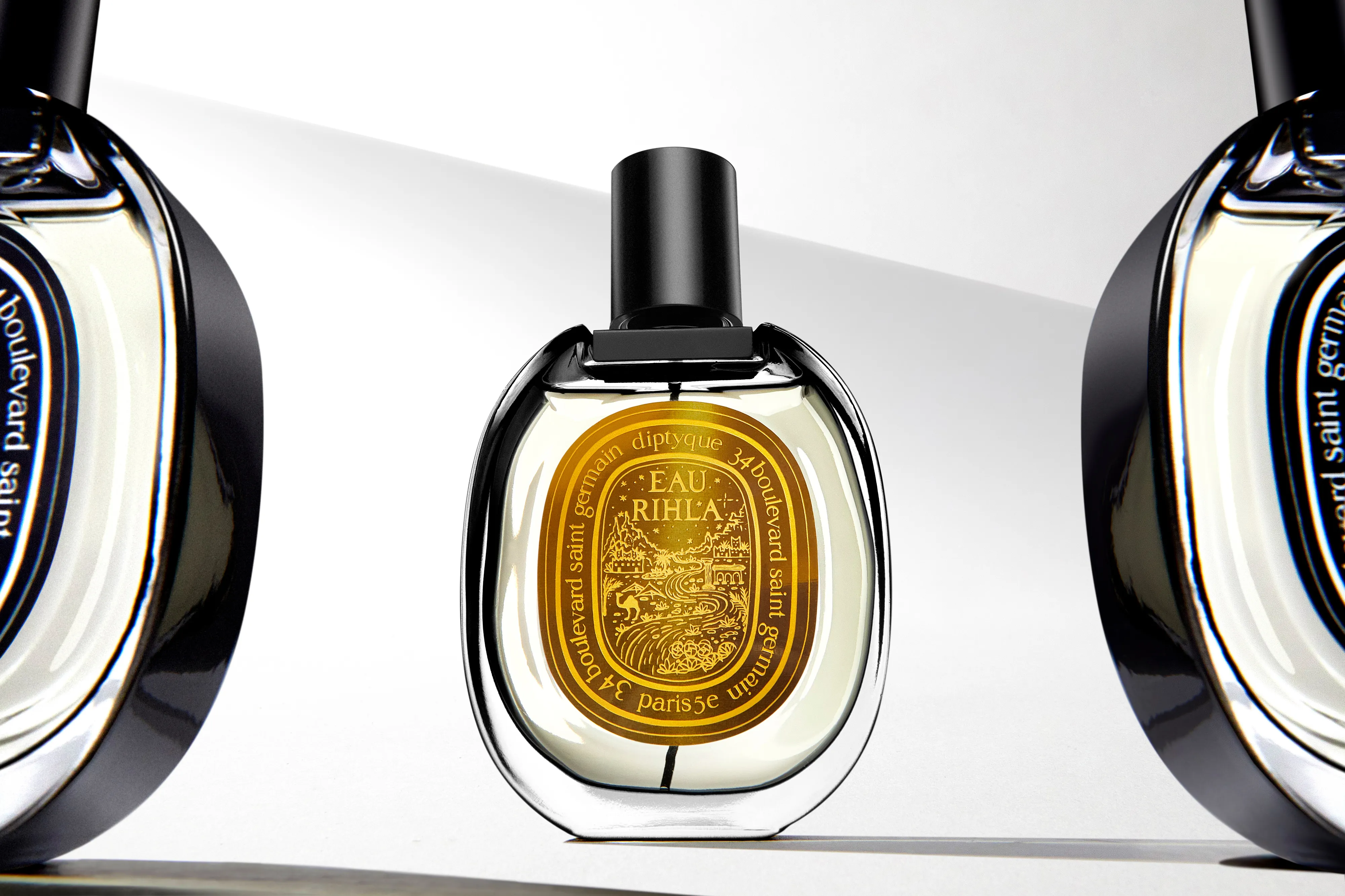 Diptyque Eau Rihla perfume bottle with a yellow label featuring a scenic design and black cap, centered with reflective bottles on each side.