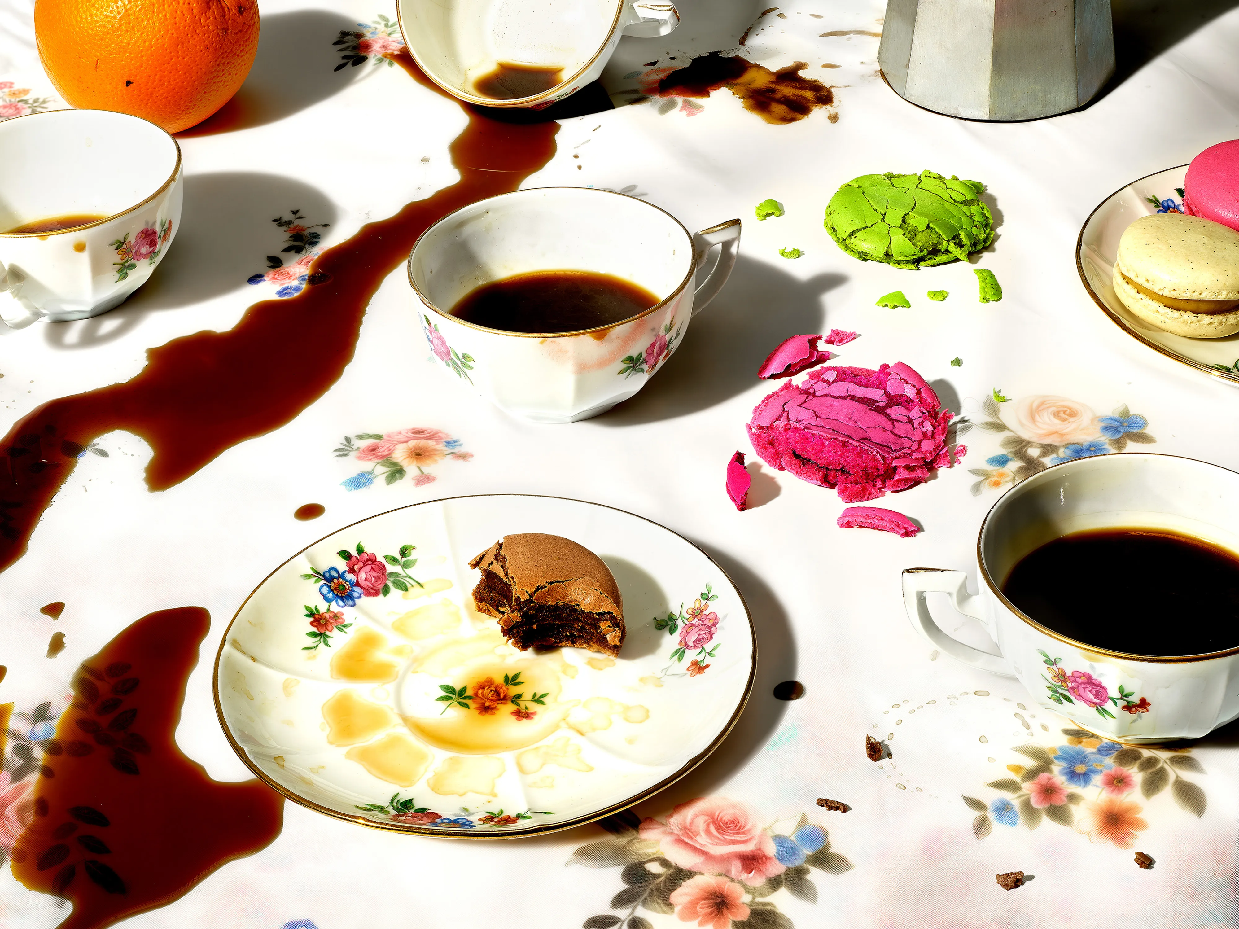 Spilled coffee across a floral tablecloth with broken macarons and floral teacups filled with coffee.
