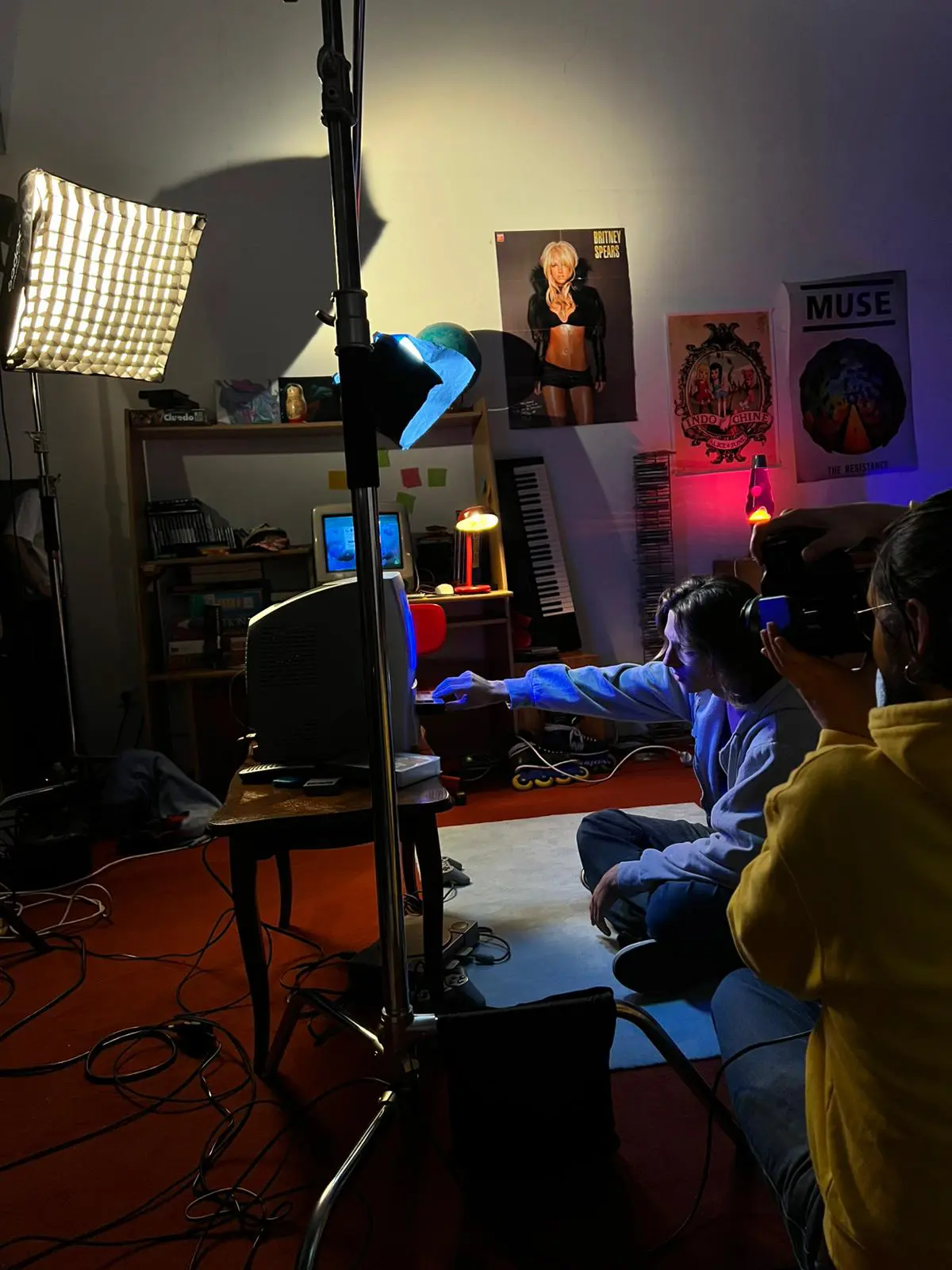 Photography session in a bedroom with one person holding a camera on the bed, another adjusting lighting, and a third viewing photos on a laptop; the room has band posters and a lava lamp.