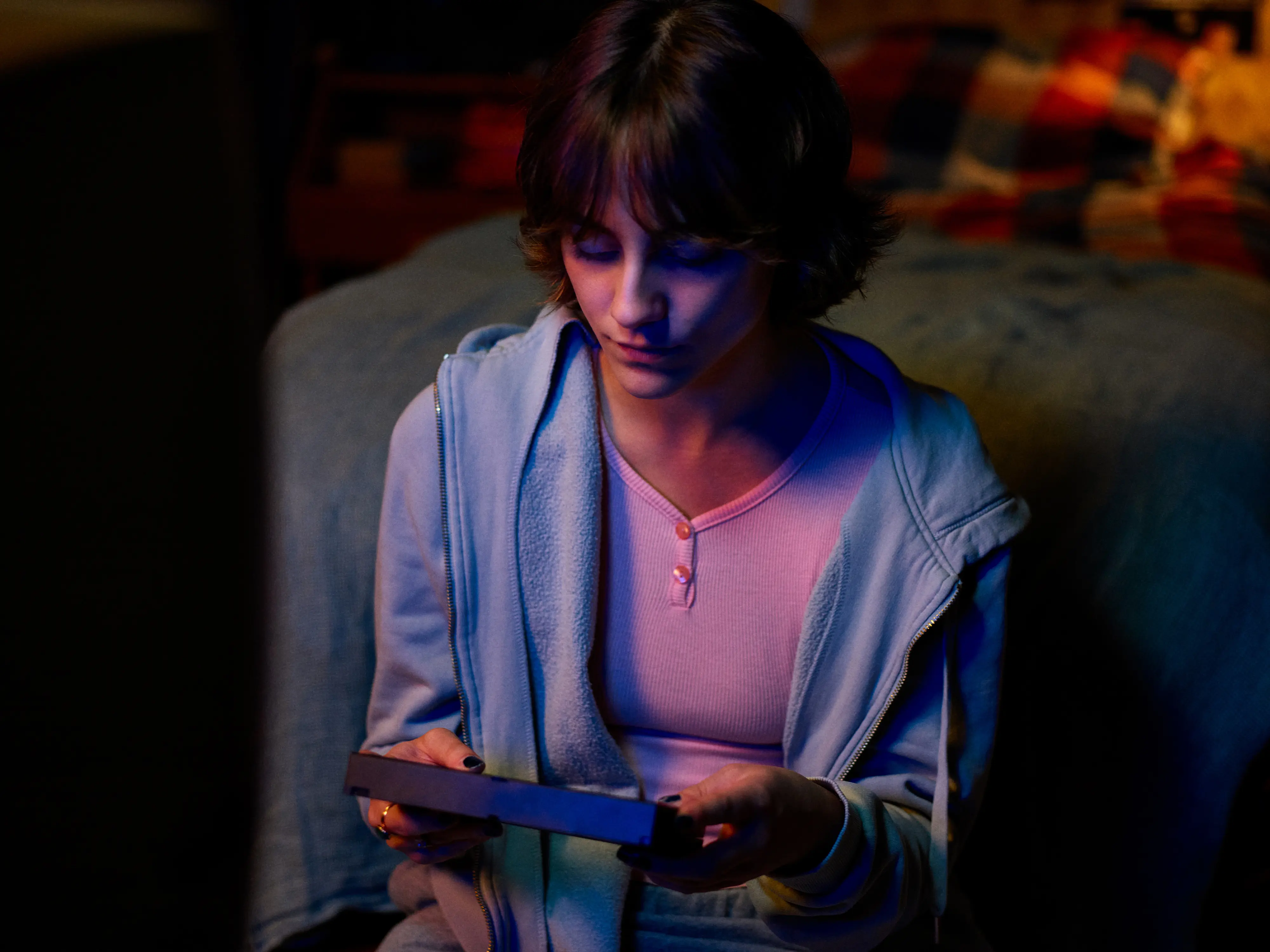 Young man with long hair illuminated by red and purple lighting looking upward thoughtfully in a dark environment.