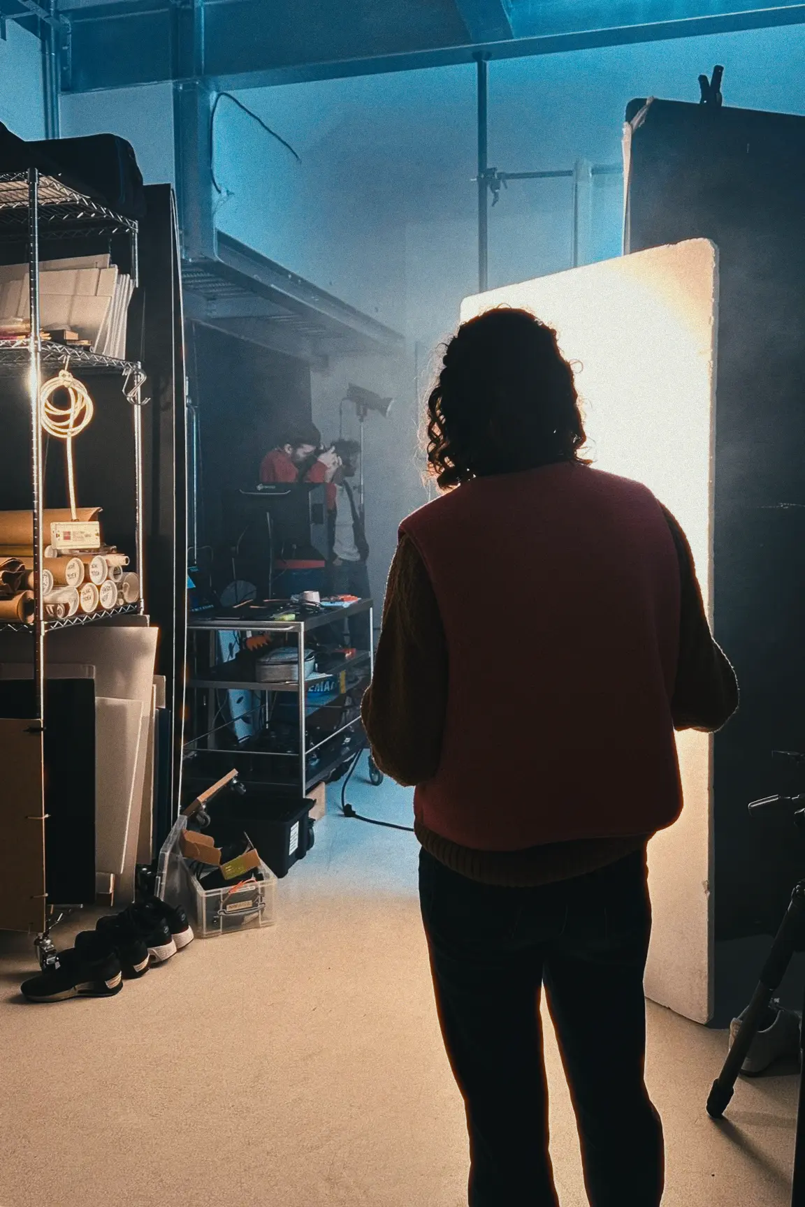Photography session in a bedroom with one person holding a camera on the bed, another adjusting lighting, and a third viewing photos on a laptop; the room has band posters and a lava lamp.