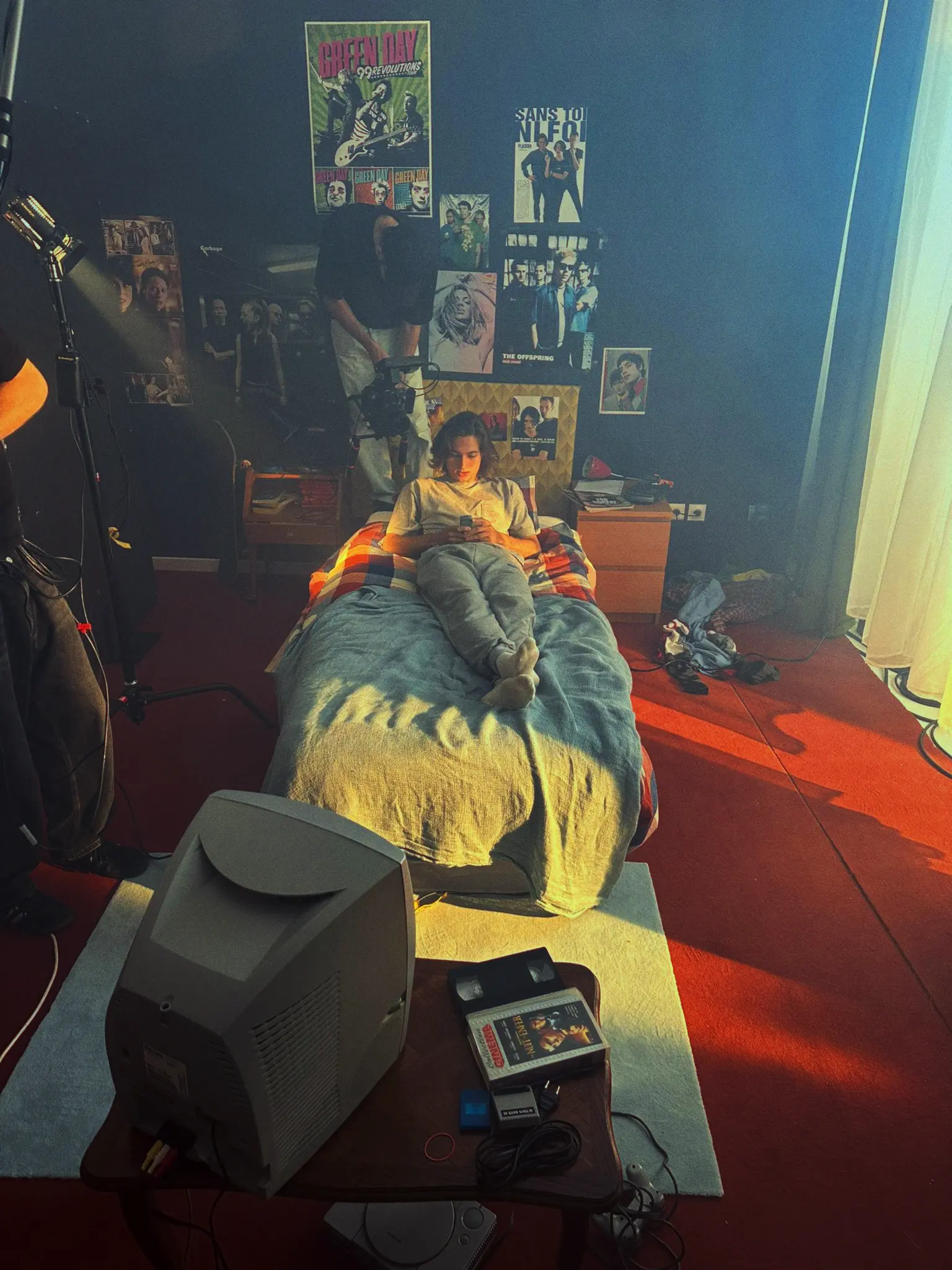 Photography session in a bedroom with one person holding a camera on the bed, another adjusting lighting, and a third viewing photos on a laptop; the room has band posters and a lava lamp.