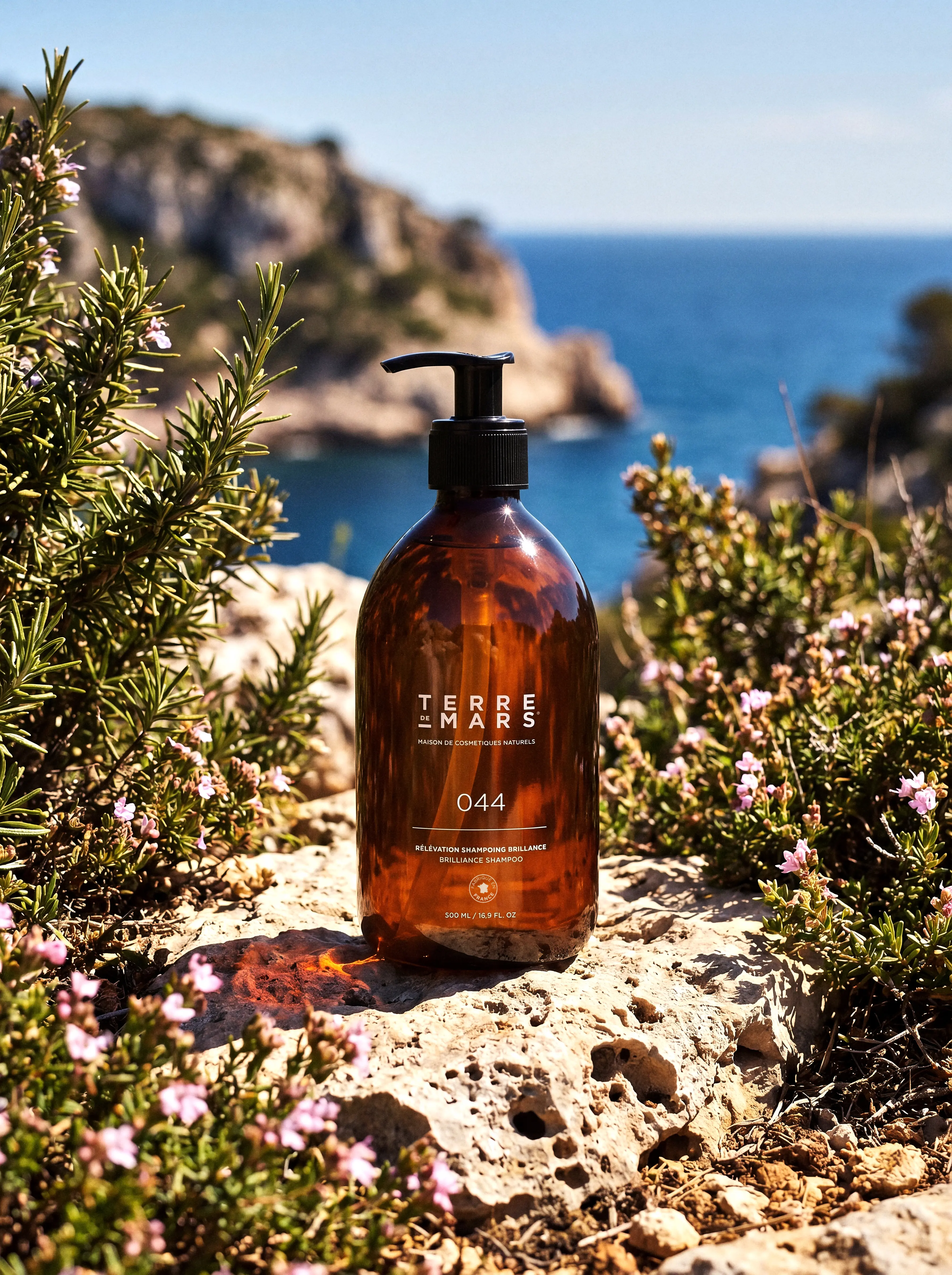 Brown Terre de Mars shampoo bottle on a rocky surface, surrounded by greenery and flowers, with sea background.