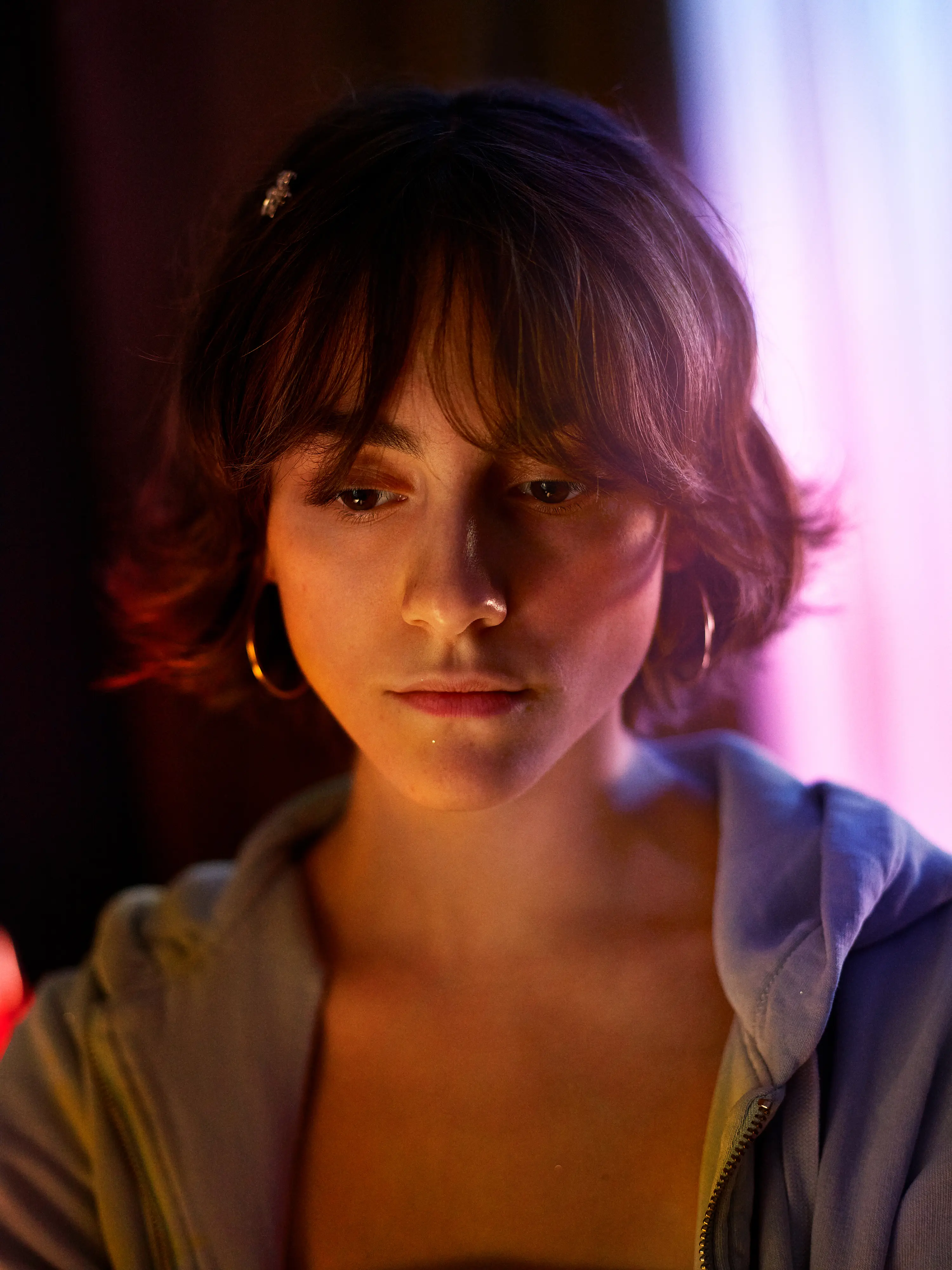 Young man with long hair illuminated by red and purple lighting looking upward thoughtfully in a dark environment.