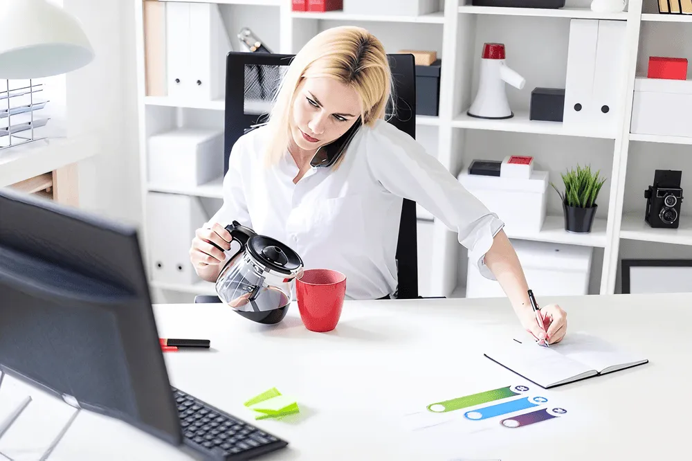 Femme blonde au bureau, parlant au téléphone tenu entre épaule et oreille, versant du café dans une tasse rouge tout en écrivant dans un carnet.