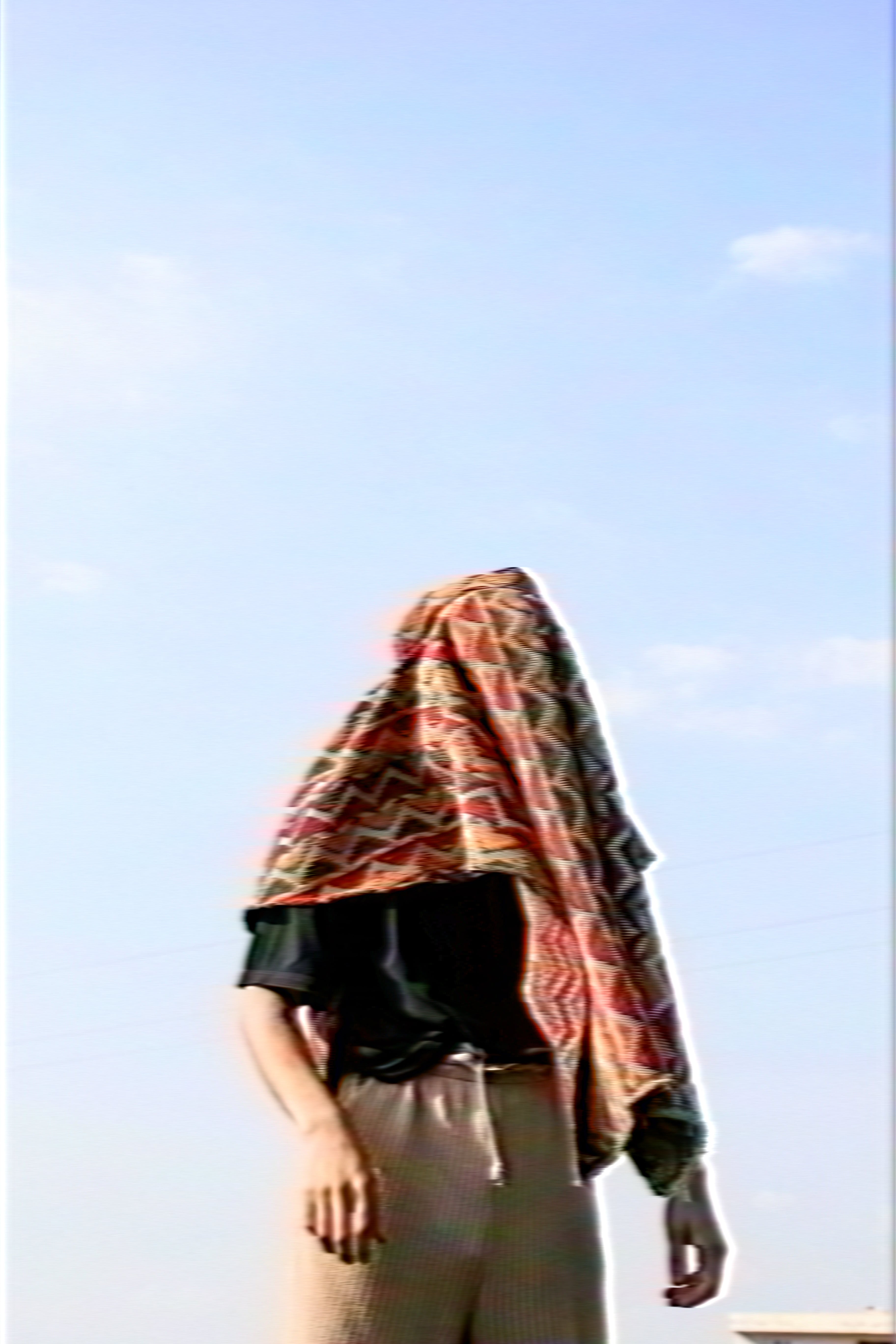 Person standing under a blue sky with a colorful patterned cloth covering their head and upper body.