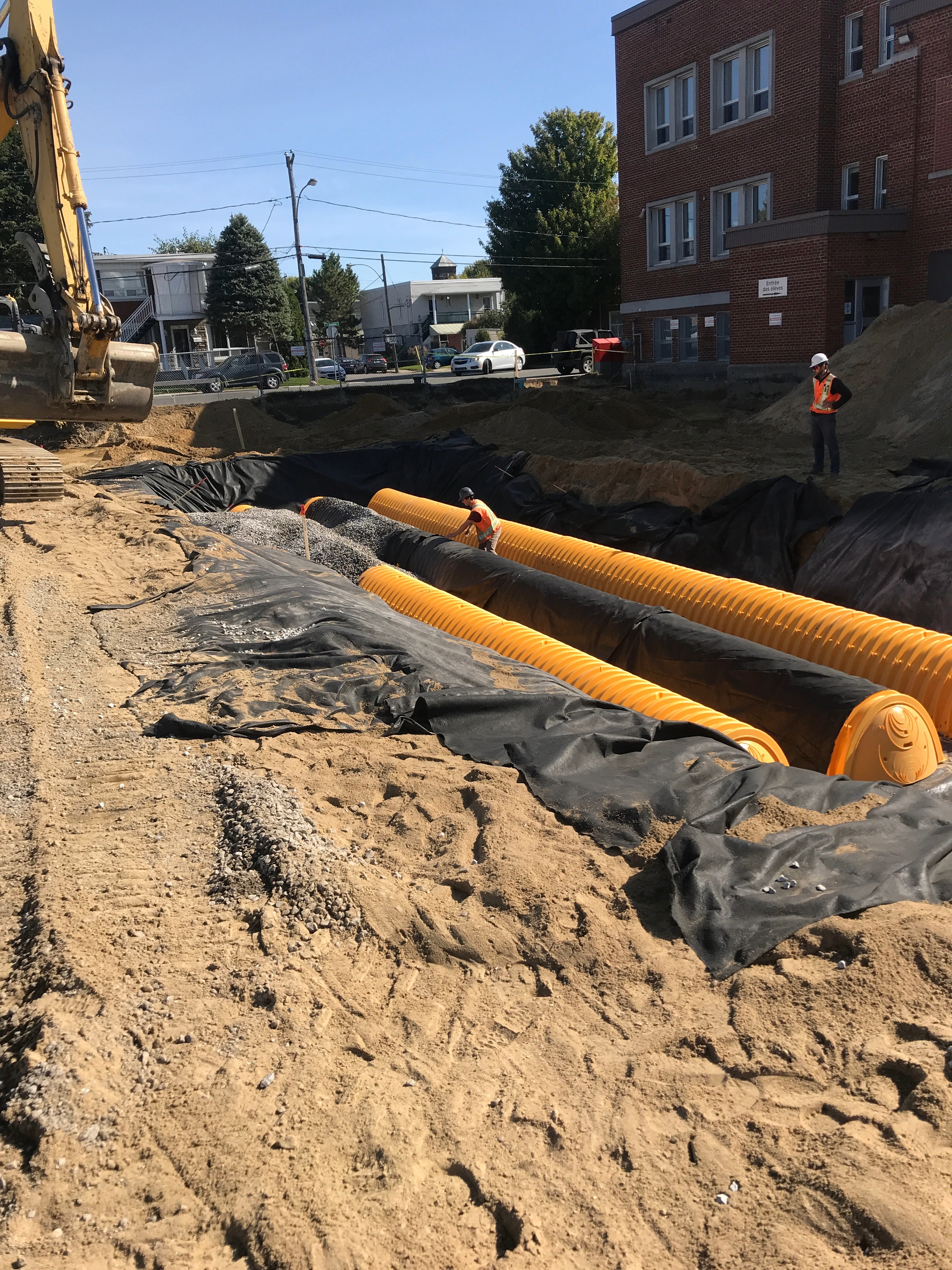 Chantier de construction avec deux travailleurs portant des casques et des gilets de sécurité orange près de grandes canalisations jaunes installées dans une tranchée recouverte de toile noire, avec une excavatrice à gauche et un bâtiment en brique à droite, en arrière-plan des arbres, des voitures et une rue.