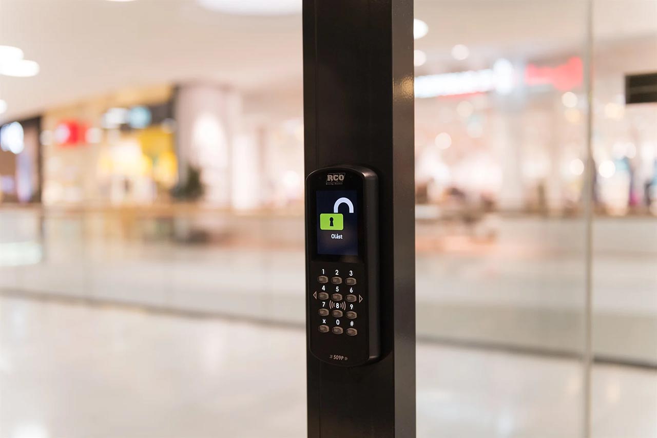 Elektronisk dørlås med talltastatur og skjerm som viser et grønt ulåst hengelåsikon med teksten "Ulåst" montert på en svart dørstolpe.