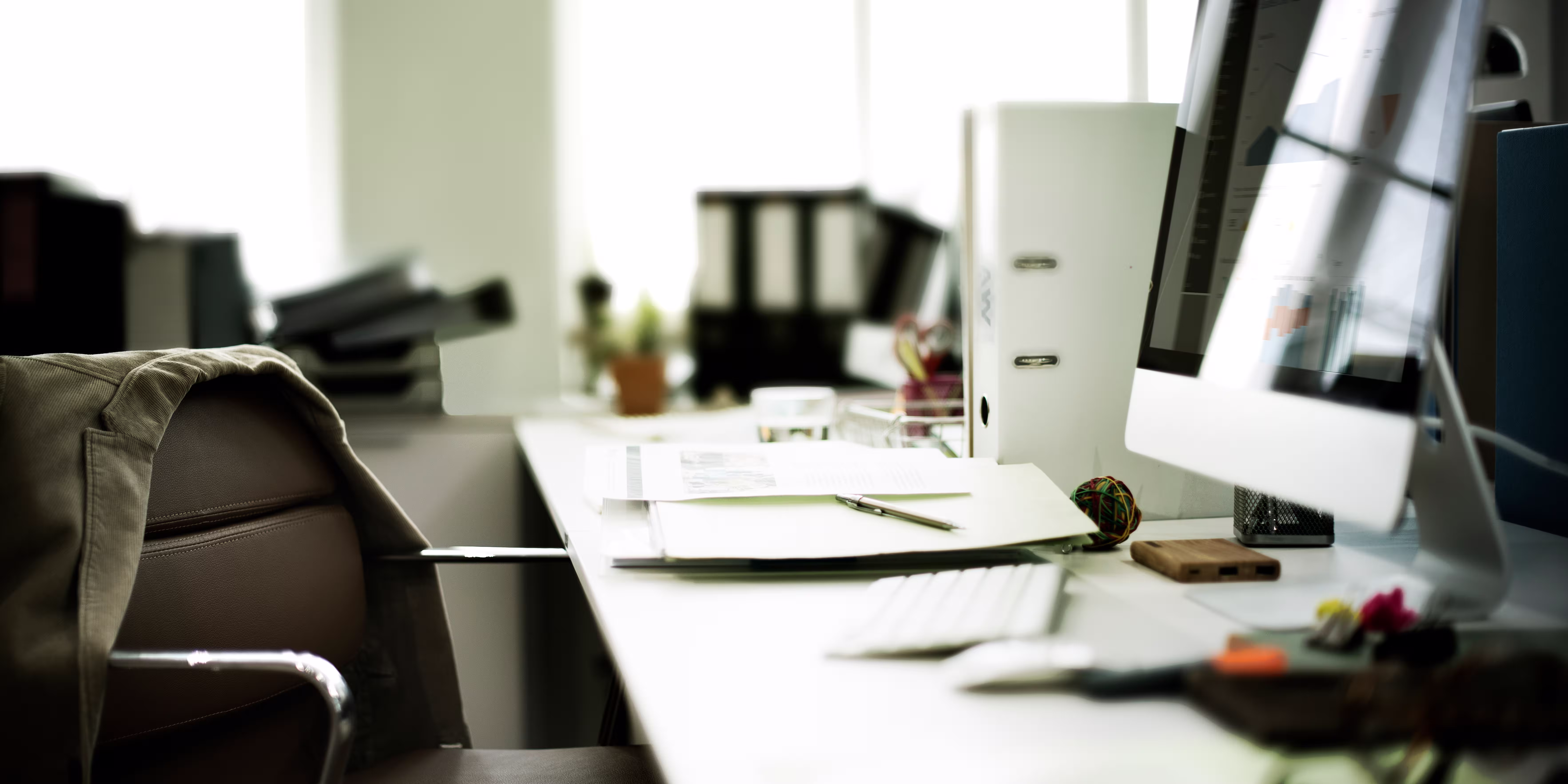 Modern office desk with computer, keyboard, files, pen, rubber band ball, and jacket draped over chair.