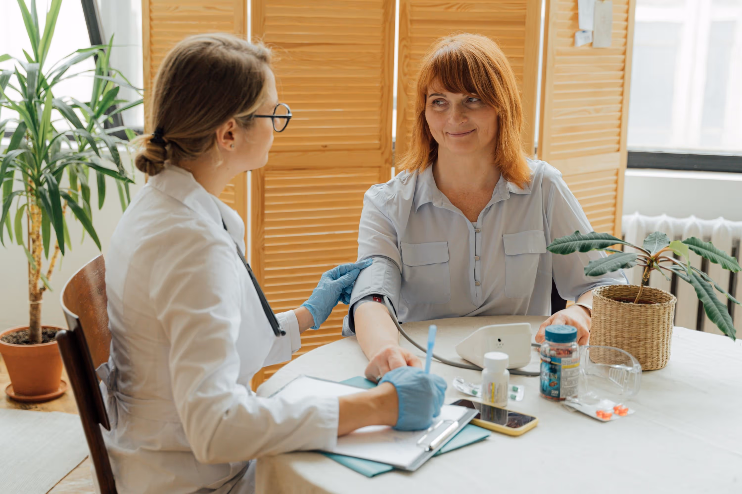 Healthcare professional in white coat measuring blood pressure of a smiling middle-aged woman at a table with medication bottles and a potted plant.