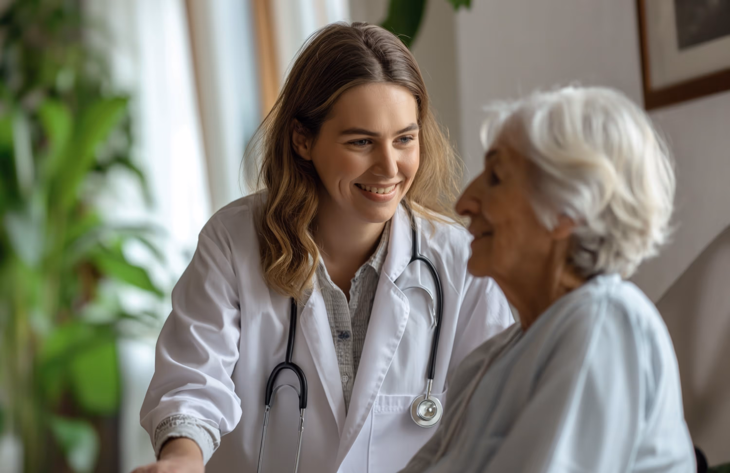 Young female doctor with a stethoscope smiling and interacting with an elderly woman in a light-filled room.
