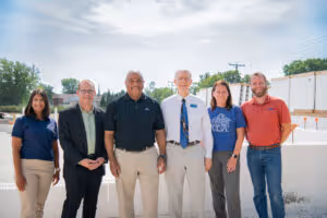 Six diverse adults standing outside in a row in casual and business casual attire against a cloudy sky background.
