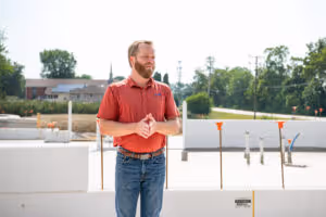 Man with a beard wearing a red polo shirt and jeans standing outdoors on a sunny day with trees and buildings in the background.