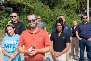 Group of eight diverse people standing outdoors, some wearing sunglasses and casual clothing, with greenery in the background.