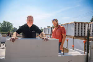 Two people working outdoors building a structure with large interlocking white blocks under a clear sky.