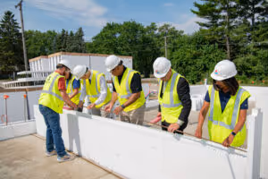 Five construction workers in safety vests and helmets installing a large white wall panel outdoors.