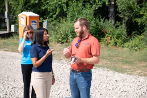Two people talking outdoors on a gravel path with a woman standing in the background near a portable toilet.