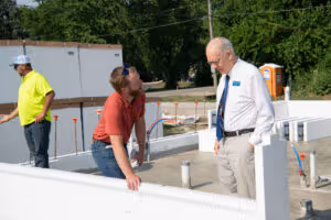 Two construction workers discussing plans inside a foundation frame with a third worker in the background.