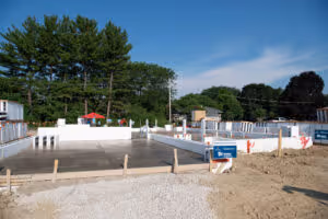 Construction site with concrete foundation and wooden framework under clear blue sky, surrounded by trees.
