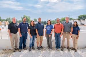 Eight people standing outdoors in a row on gravel with a fence and sky in the background.