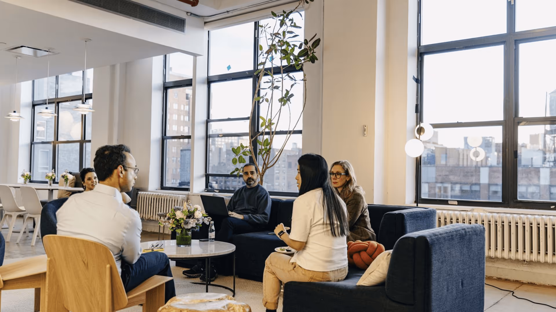 Group of five people having a discussion in a modern office lounge with large windows and natural light.