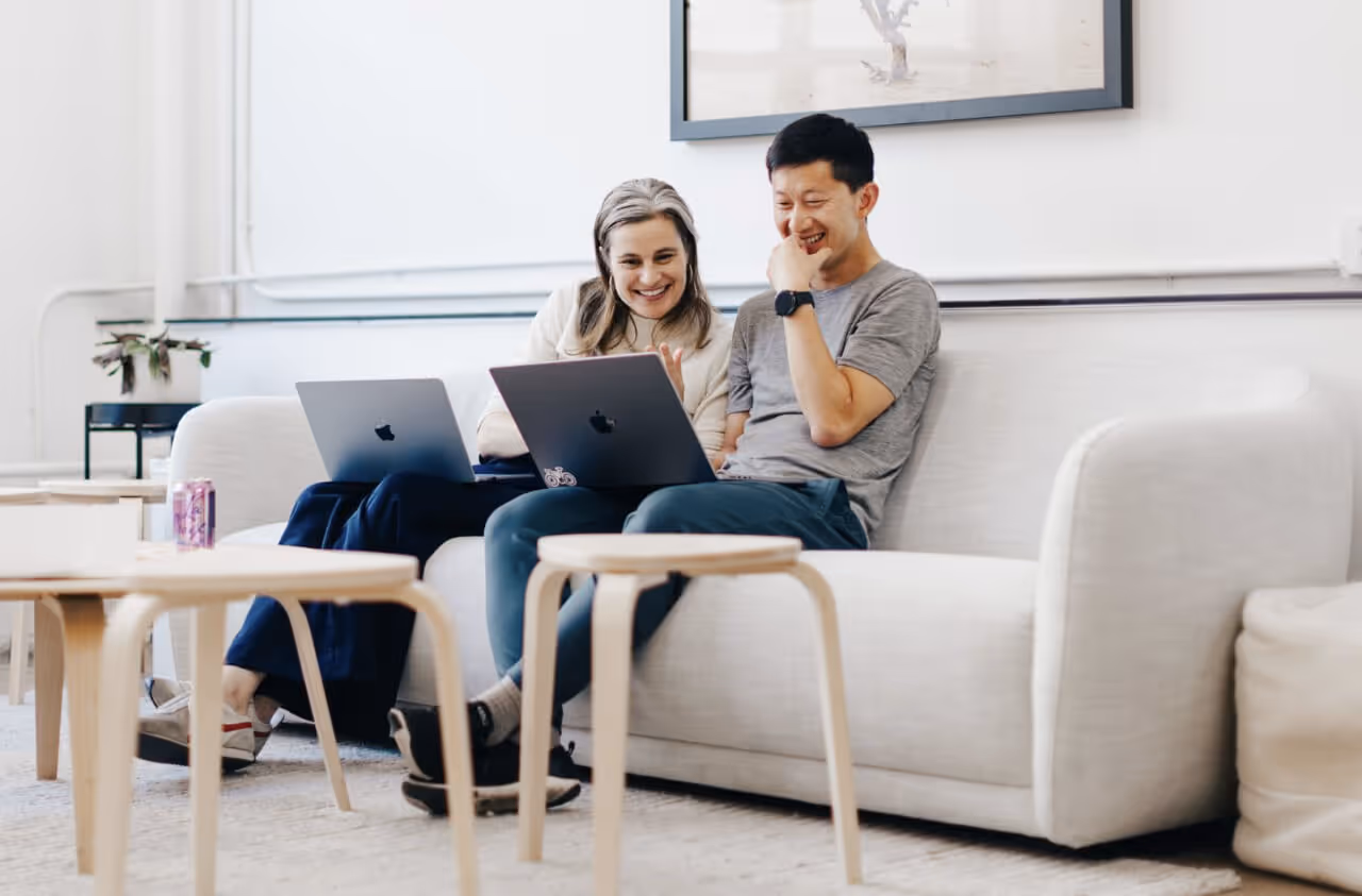 Two coworkers sit on a couch, smiling at laptops in a bright office