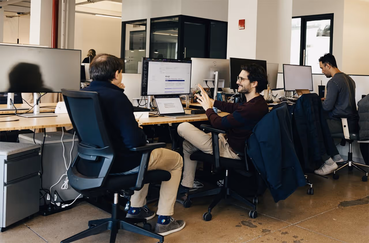 Two coworkers talk at computer desks in an open-plan office
