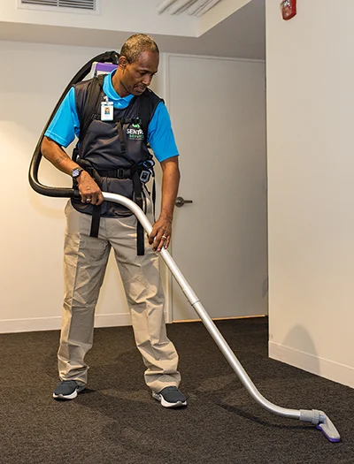 A man wearing a blue shirt and black vest cleaning a carpeted floor with a professional vacuum cleaner inside a room.