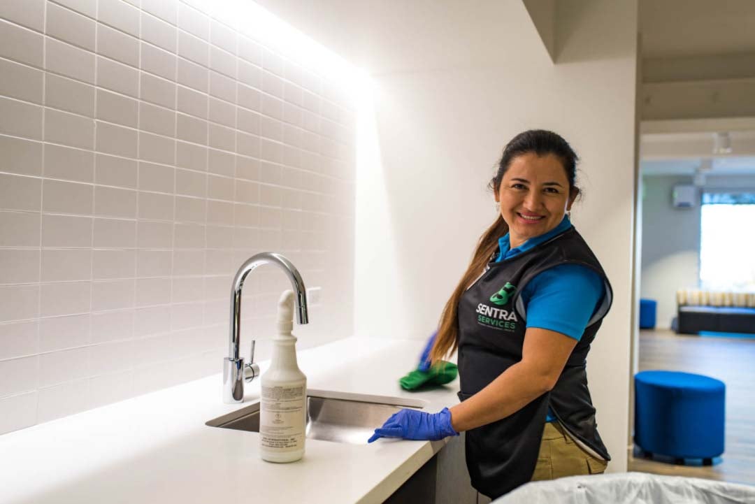 A person cleaning a shiny floor in a room with dark stools lined up along a counter and tables with chairs by windows with blinds.