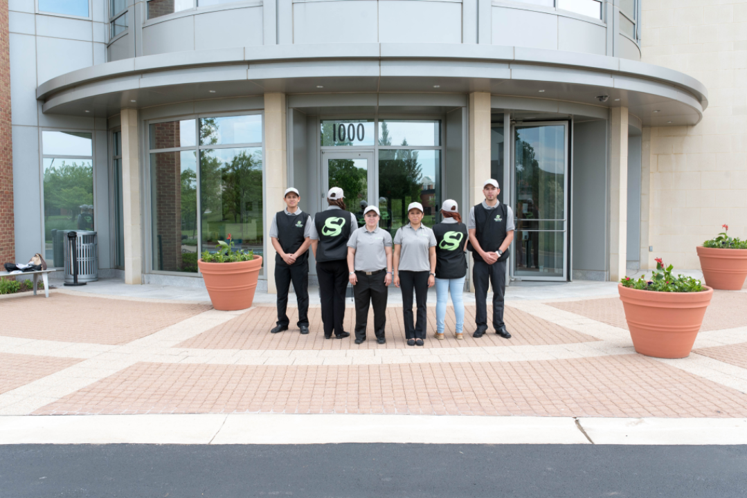 Professional cleaner wearing gloves and uniform wiping a large public restroom sink with a yellow cloth, with cleaning supplies cart nearby.
