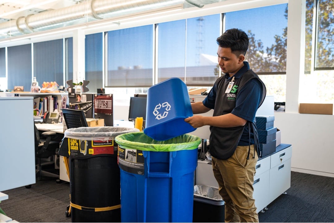 A person cleaning a shiny floor in a room with dark stools lined up along a counter and tables with chairs by windows with blinds.