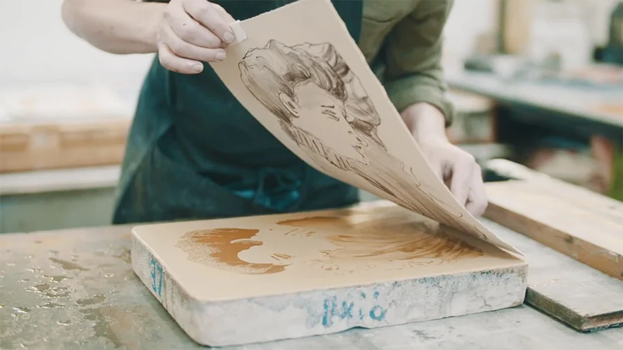 A printmaker carefully lifts a sheet of paper from a lithography stone, revealing a freshly transferred image.